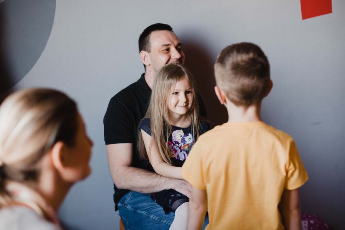 Mężczyzna uśmiecha się, podczas gdy młoda dziewczyna siedzi na jego kolanach. Naprzeciw nich stoi chłopiec w żółtej koszuli, a na pierwszym planie częściowo widoczna jest kobieta odwrócona tyłem do kamery. Ta interakcja sugeruje szczęśliwe i zrelaksowane rodzinne spotkanie, doskonale uchwycone w tej chwili na fotografii eventowej.