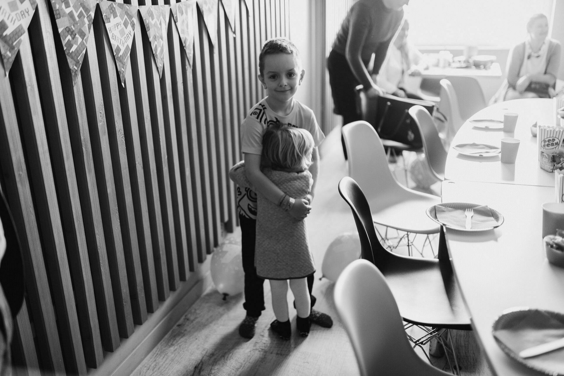 Black and white photo of two children hugging at a birthday party. They are standing at a long table decorated with party decorations, plates and cups. In the background you can see several adults and children engaged in various activities - an excellent event photo by event photographer Warsaw.  