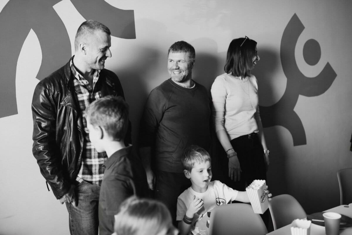 Czarno-białe zdjęcie trójki dorosłych i trójki dzieci stojących razem, uśmiechniętych i najwyraźniej pogrążonych w rozmowie. Jedno dziecko trzyma pojemnik z popcornem. Tło przedstawia abstrakcyjną grafikę ścienną, utrwalającą idealny moment w fotorelacji z wydarzeń autorstwa fotografa eventowego Warszawa.