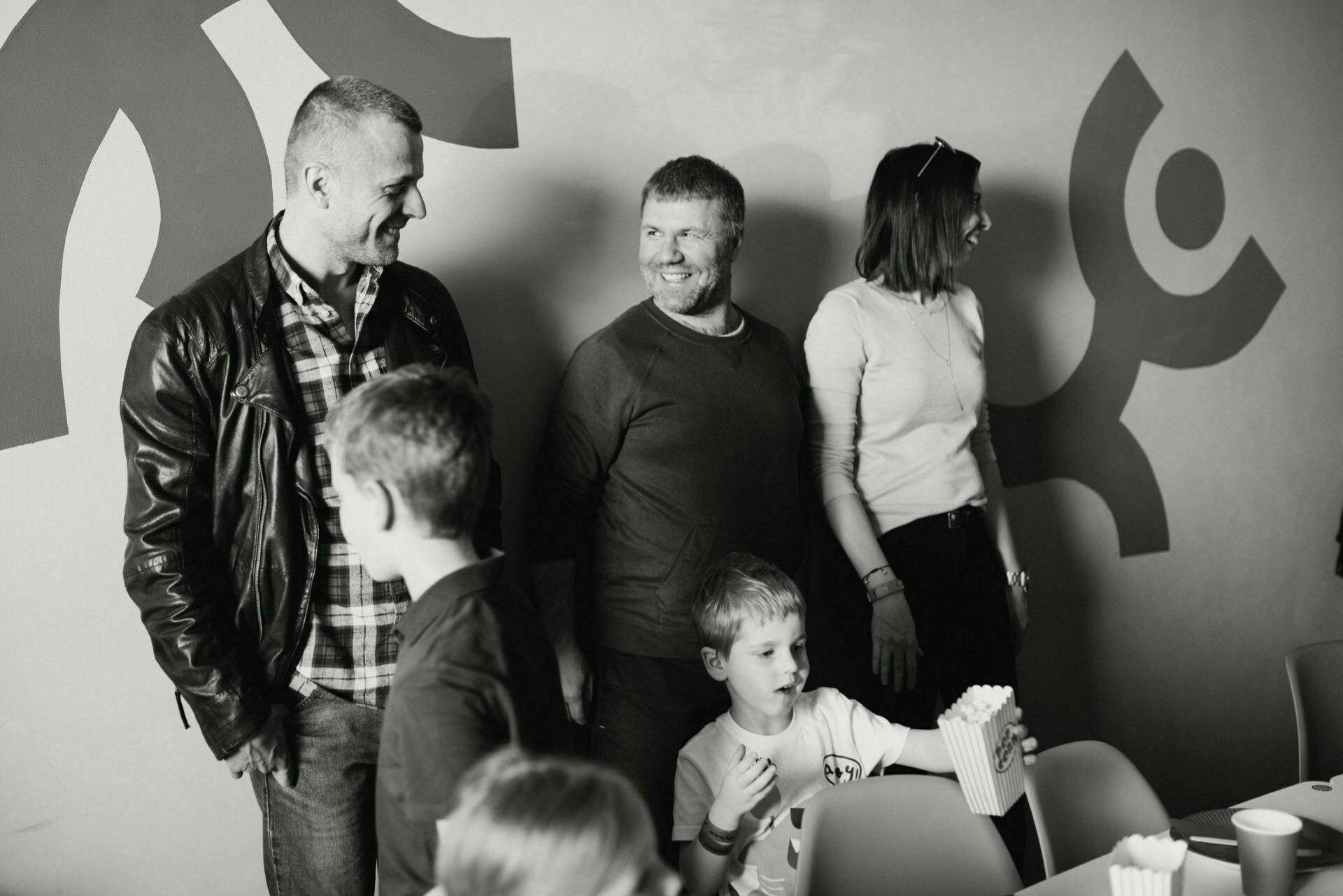 Black and white photo of three adults and three children standing together, smiling and apparently immersed in conversation. One child is holding a container of popcorn. The background depicts an abstract wall graphic, capturing the perfect moment in an event photo essay by event photographer Warsaw.  