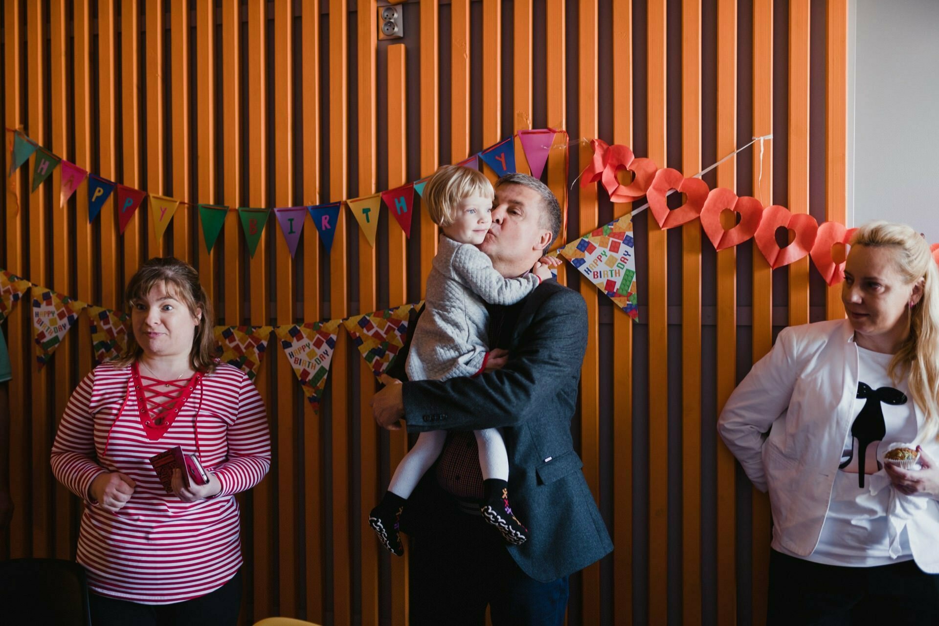 A man holding and kissing a baby stands in front of a colorful "Happy Birthday" banner. On the left looks a woman in a red and white striped shirt, on the right stands another woman in a white jacket and black tie, all against a background of wooden panels - beautifully captured by an event photographer in Warsaw. 