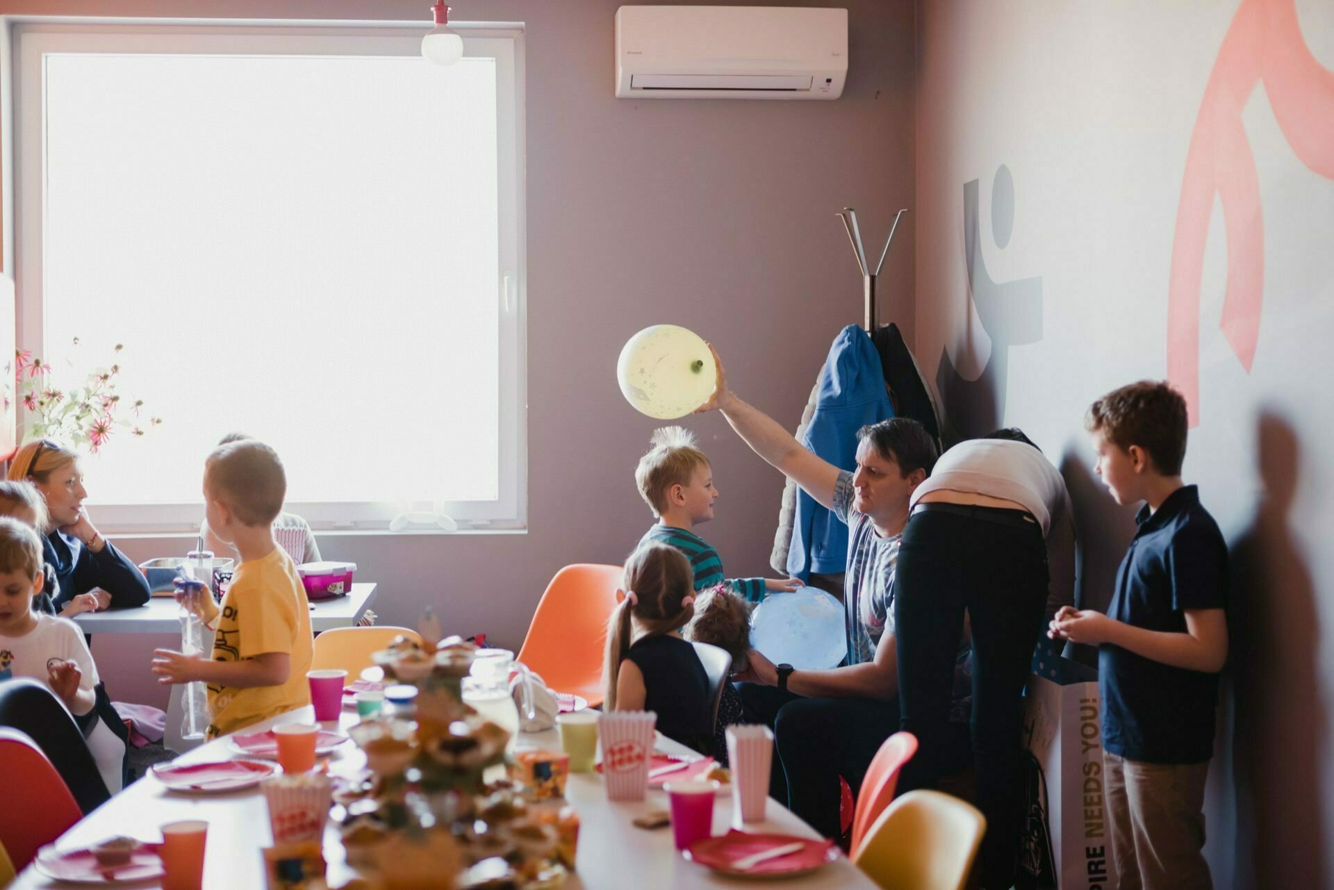 A lively children's party where children gathered around a table decorated with plates, cups and snacks. Adults help the children with activities. A person inflates a balloon near the window while others are busy talking. The room is colorful and festive, perfect for a memorable photo-op of the events.   