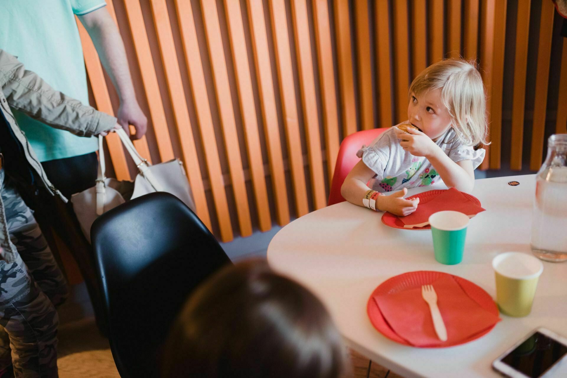 A small child with blond hair sits at a round table eating a snack. On the table are red paper plates, a green cup, a white cup and a glass decanter. An adult and another person stand nearby. In the background are wooden vertical slats that capture the moment perfect for a photo event.   