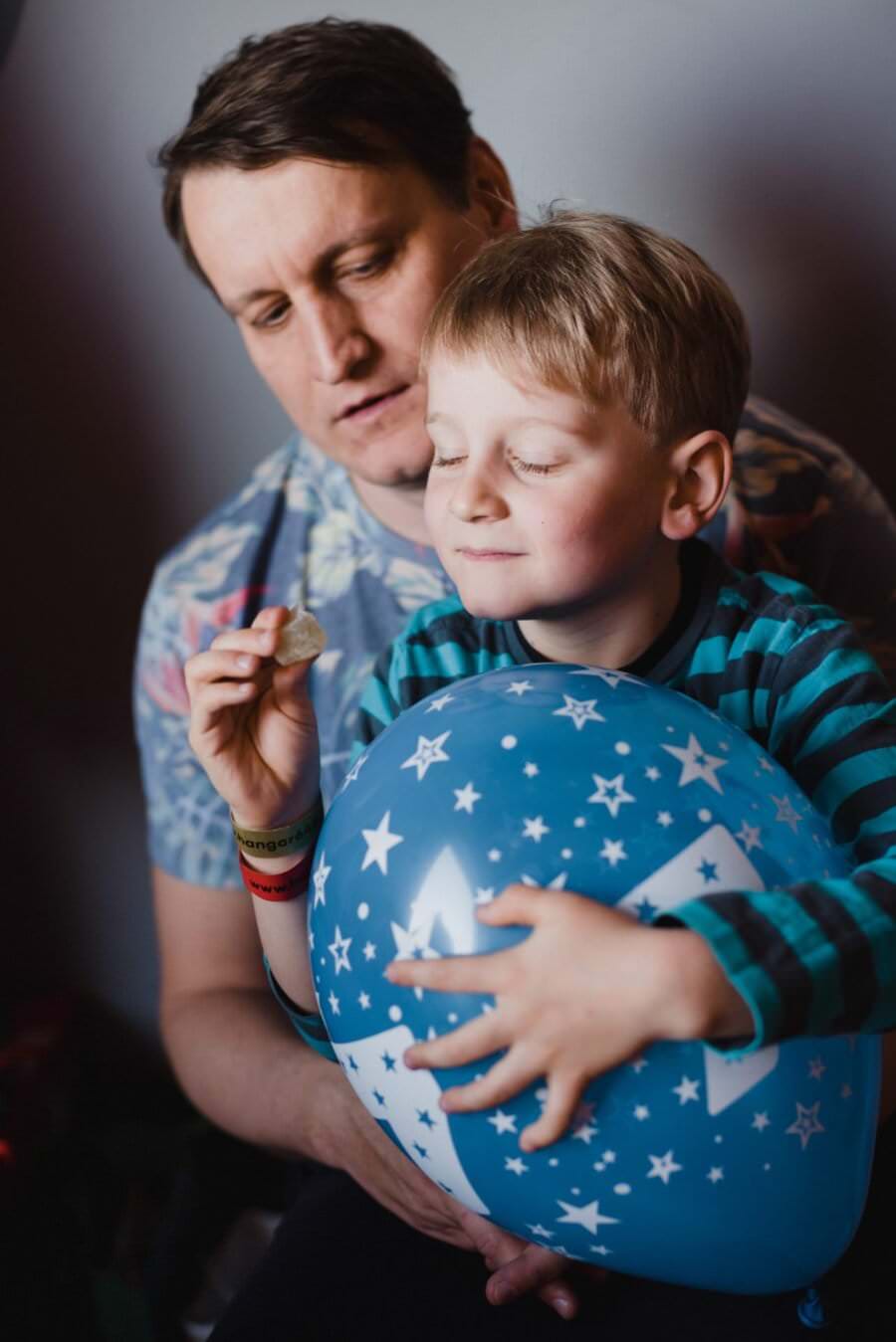 Uśmiechnięty mężczyzna i młody chłopak. Chłopiec trzyma niebieski balonik z białymi wzorami gwiazdek i ma zamknięte oczy, trzymając mały przedmiot. Stojący za nim mężczyzna o łagodnym wyrazie twarzy zdaje się być doskonale uchwycony w tym przykładzie fotografii eventowej.