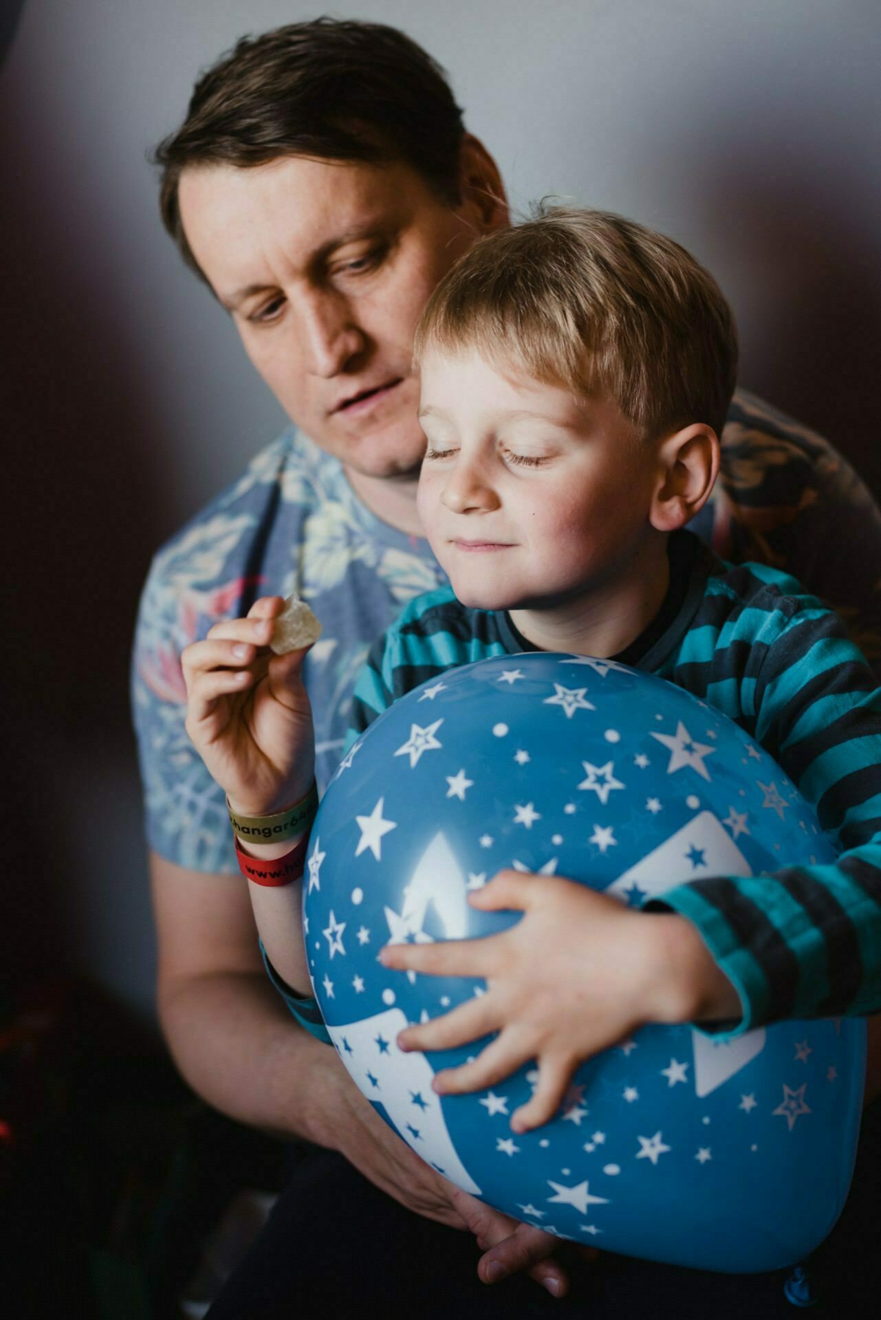 A smiling man and a young boy. The boy is holding a blue balloon with white star patterns and has his eyes closed, holding a small object. The man standing behind him with a gentle expression seems to be perfectly captured in this example of event photography.  