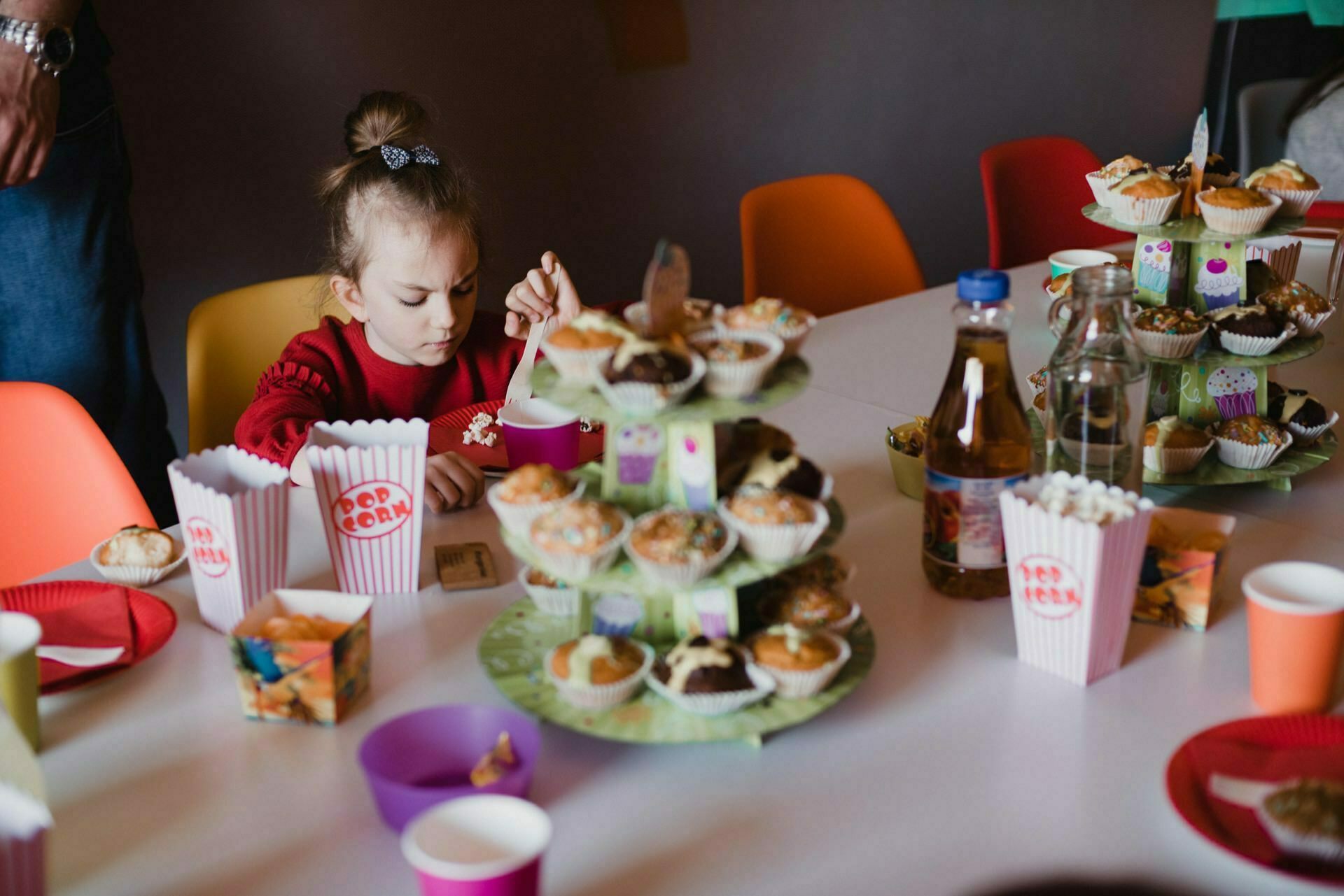 A young girl in a red shirt sits at a table decorated for the party, surrounded by various cupcakes, paper cups and colorful plates. The table also contains popcorn, drinks and other snacks. In the background you can see brightly colored chairs - a perfect scene for event photography.  