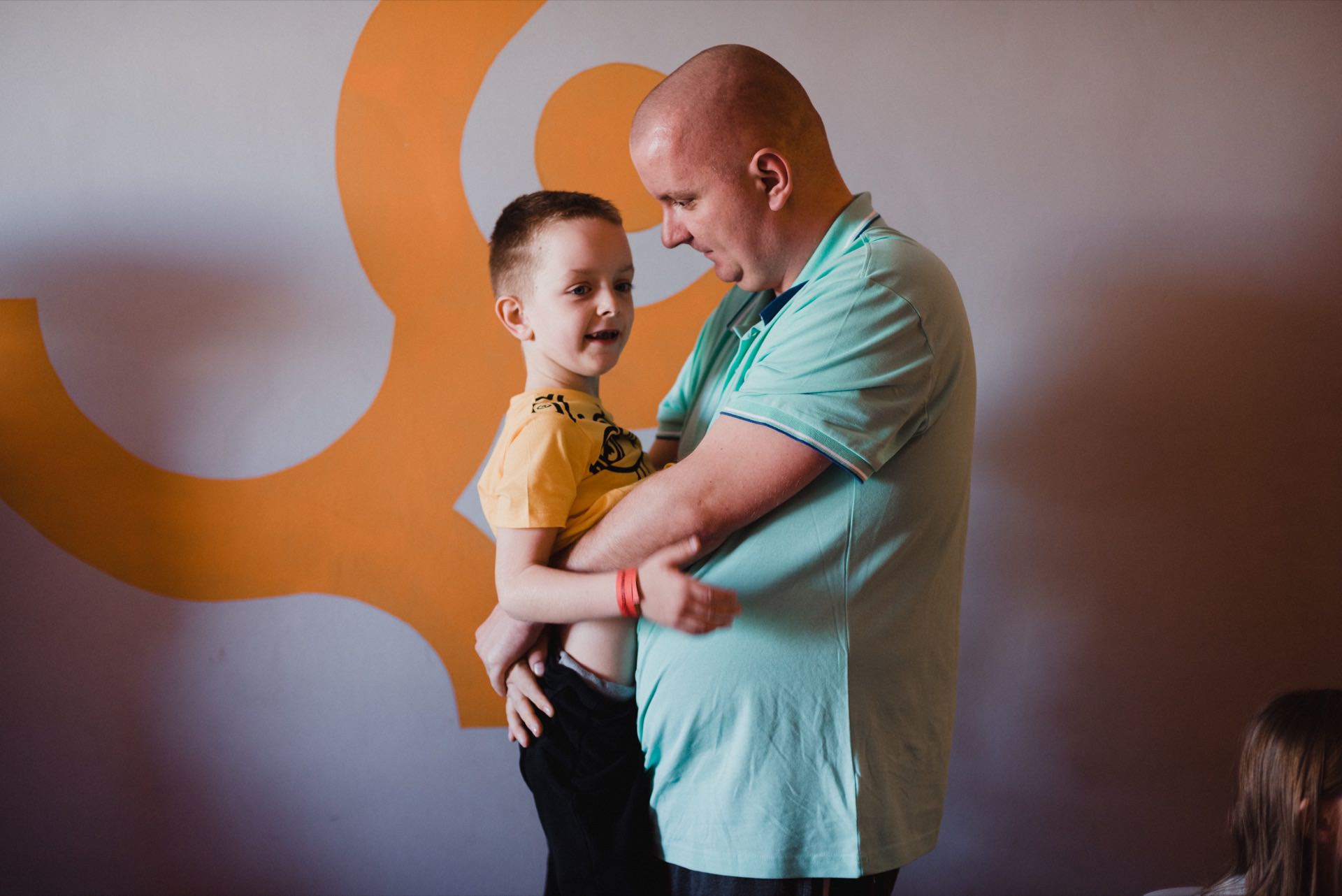 A bald man in a light blue polo shirt is holding a young boy in a yellow shirt. They are in a room, and the wall behind them depicts a large orange spiral. The two appear to be smiling at each other, sharing a tender moment perfectly captured by event photographer Warsaw.  