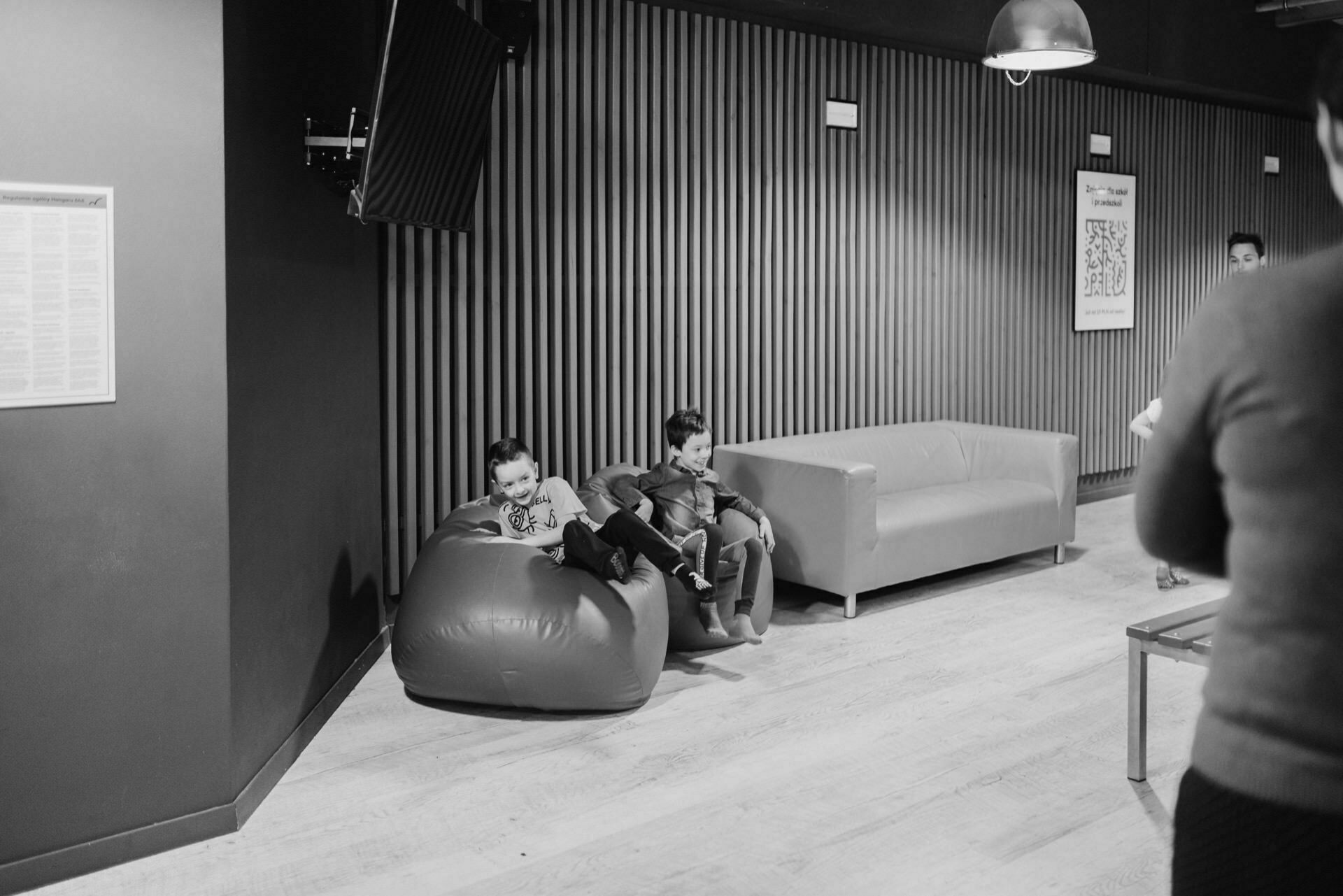 The monochrome photo shows two children resting on beanbags in a living room with a couch and TV mounted on the wall. An adult partially enters the frame on the right. The space, ideal for a photo event, features a modern, cozy decor with wooden floors and vertical wall panels.  