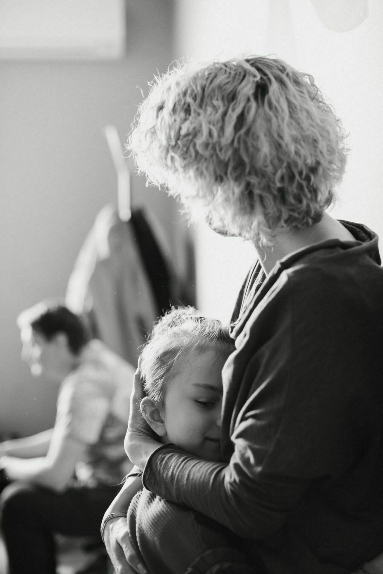 The black-and-white photo of the event shows a woman with curly hair holding a small child, who is resting his head on her chest with a calm expression on his face. In the background, a person can be seen sitting and concentrating on some activity. The scene is delicate and intimate.  