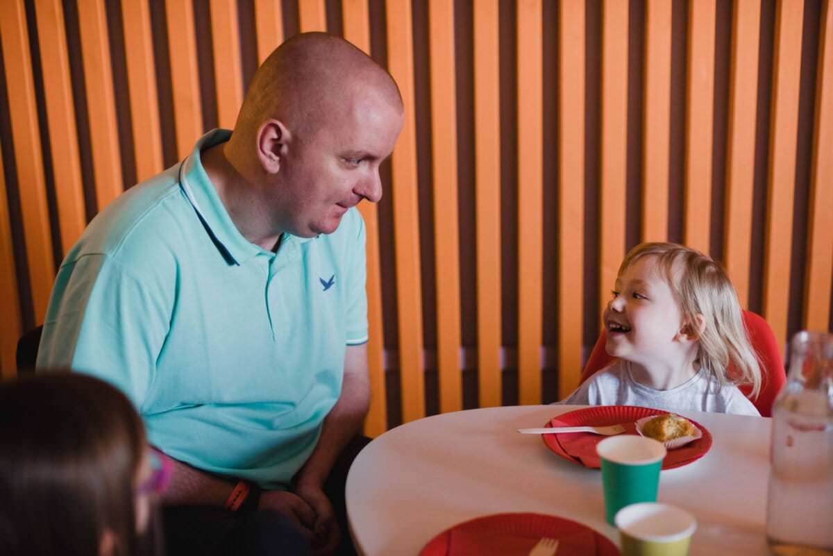 Mężczyzna w jasnoniebieskiej koszuli siedzi przy stole i uśmiecha się do małej dziewczynki o blond włosach, która również uśmiecha się do niego. Dzielą się posiłkiem, a na stole stoją talerze i kubki. W tle drewniana boazeria – idealna scena do fotografii eventowej.