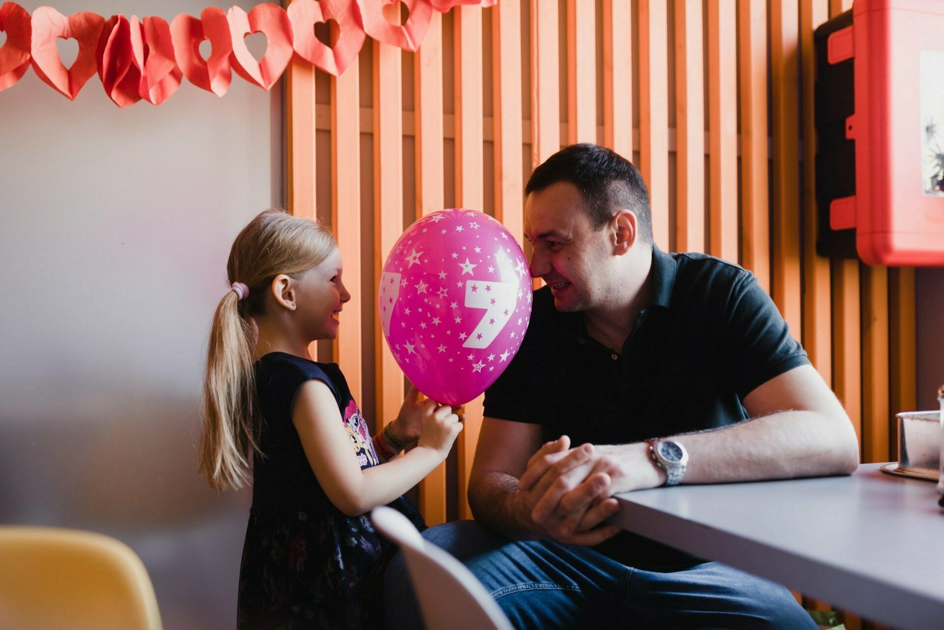 A young girl with a ponytail holds a pink balloon with stars and the number 7, smiling at a seated man in a black shirt. They are in a room decorated with a garland of red hearts on the wall. The man is also smiling, leaning over the table, which was perfectly captured by an event photographer Warsaw.  