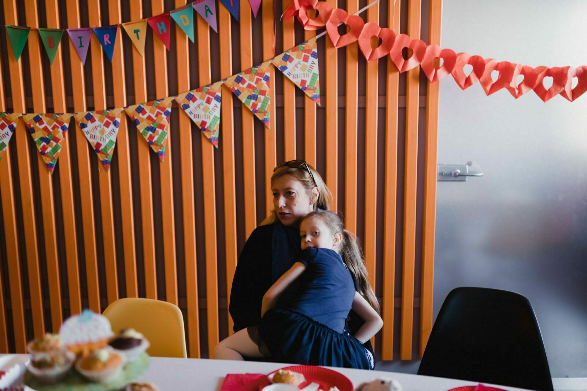 A woman sits and hugs a young girl in front of a wooden wall lined with paneling decorated with colorful banners reading "Happy Birthday" and heart-shaped paper chains. In the foreground is a table with various cupcakes and cakes. The atmosphere is festive and warm, capturing the perfect photo report of events filled with joy.  