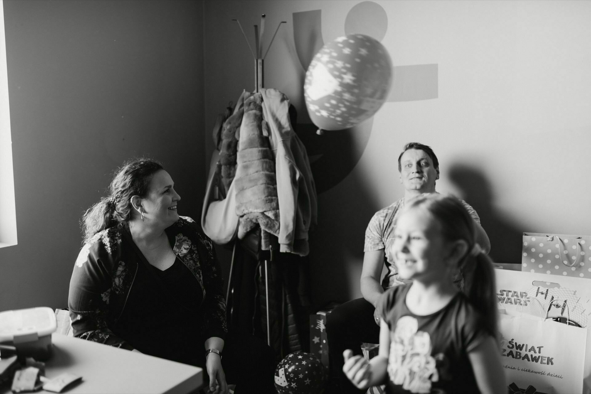 The black-and-white photo captures a joyful moment of the event. A woman, a man and a child are sitting in a room with a coat rack and various objects. The child is holding a balloon, while the adults look on with smiles on their faces. Luggage and clutter create an authentic backdrop for this event photography.   