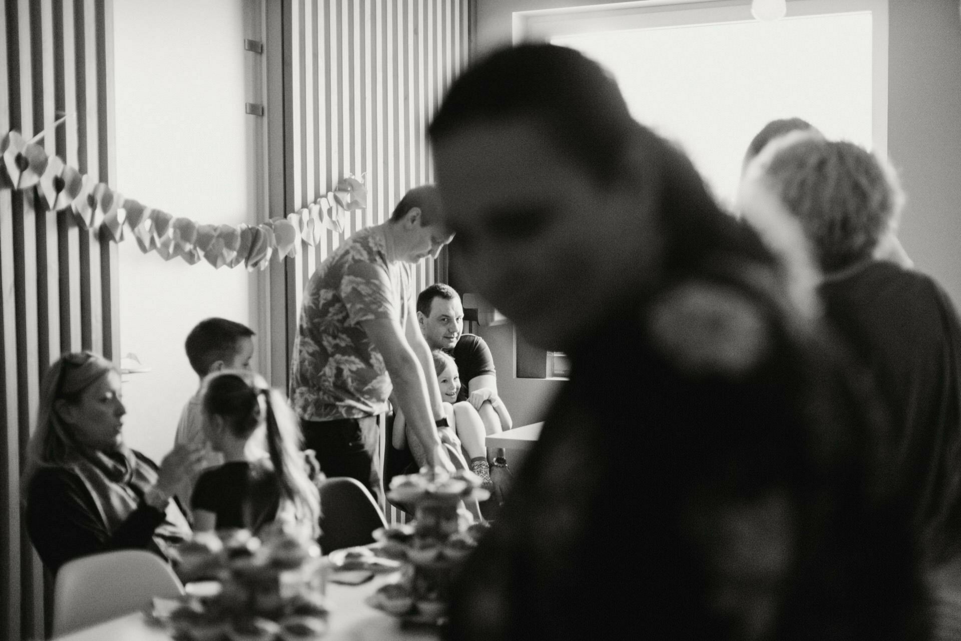   Black and white image of a busy gathering in a room. Some people sit at a table with stacks of trays of goodies, while others stand. Paper decorations hang on the wall. The foreground is out of focus, and the figure in the background catches the eye - a perfect photo-reportage of the events.   