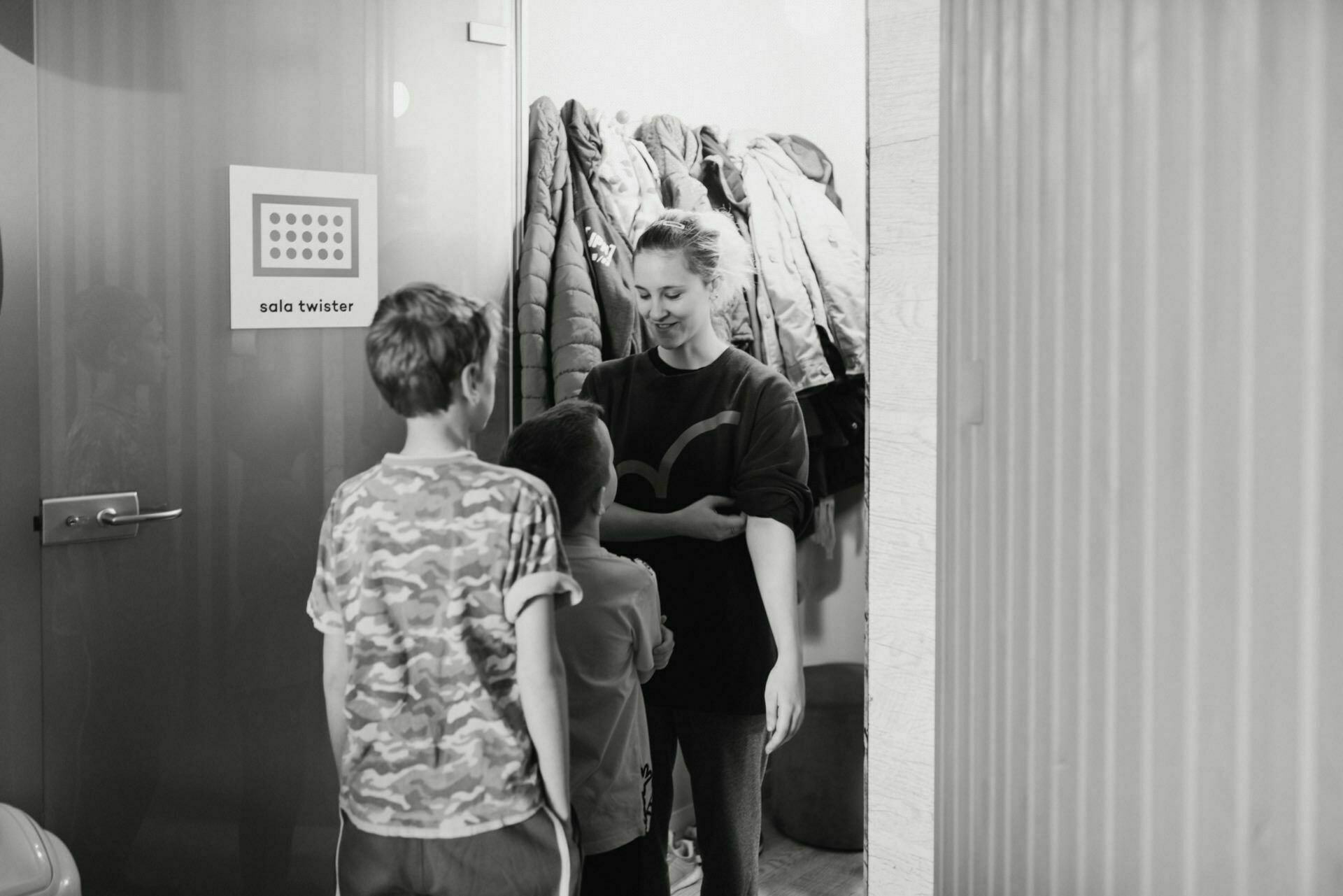 The grayscale painting shows a woman and two children standing near a door. The woman smiles, looks at one child and corrects her sleeve. The children stand facing her, one wearing a camouflage shirt. In the background, coats are hanging on hangers - a perfect example of event photography captured by an expert event photographer warsaw.   