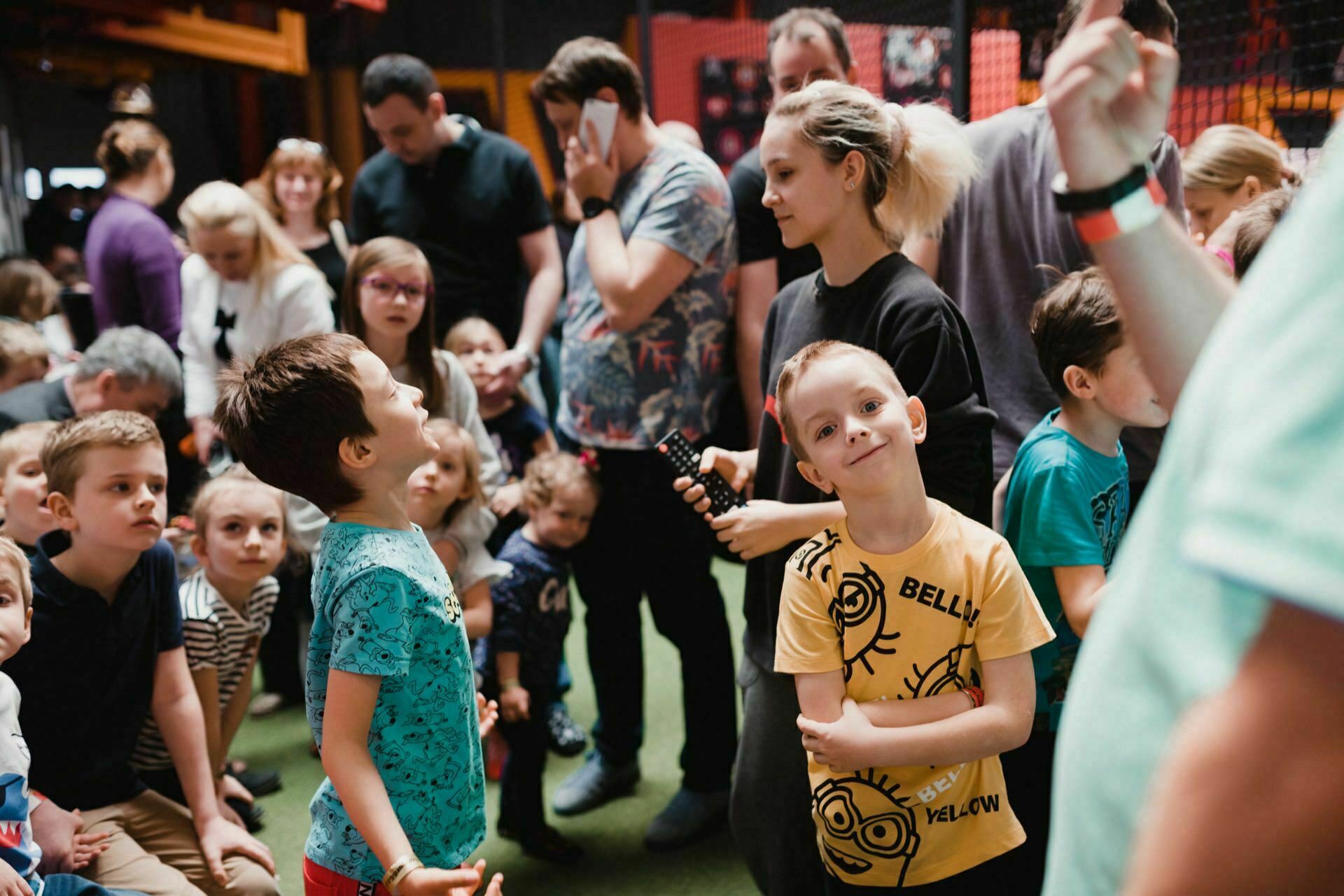 A lively group of children and adults have gathered in the room. Some children stand while others sit on the floor, engaging in various activities. Several adults look after the children, capturing moments in a photo essay of the event. The setting appears to be a playground or an event in the room.   