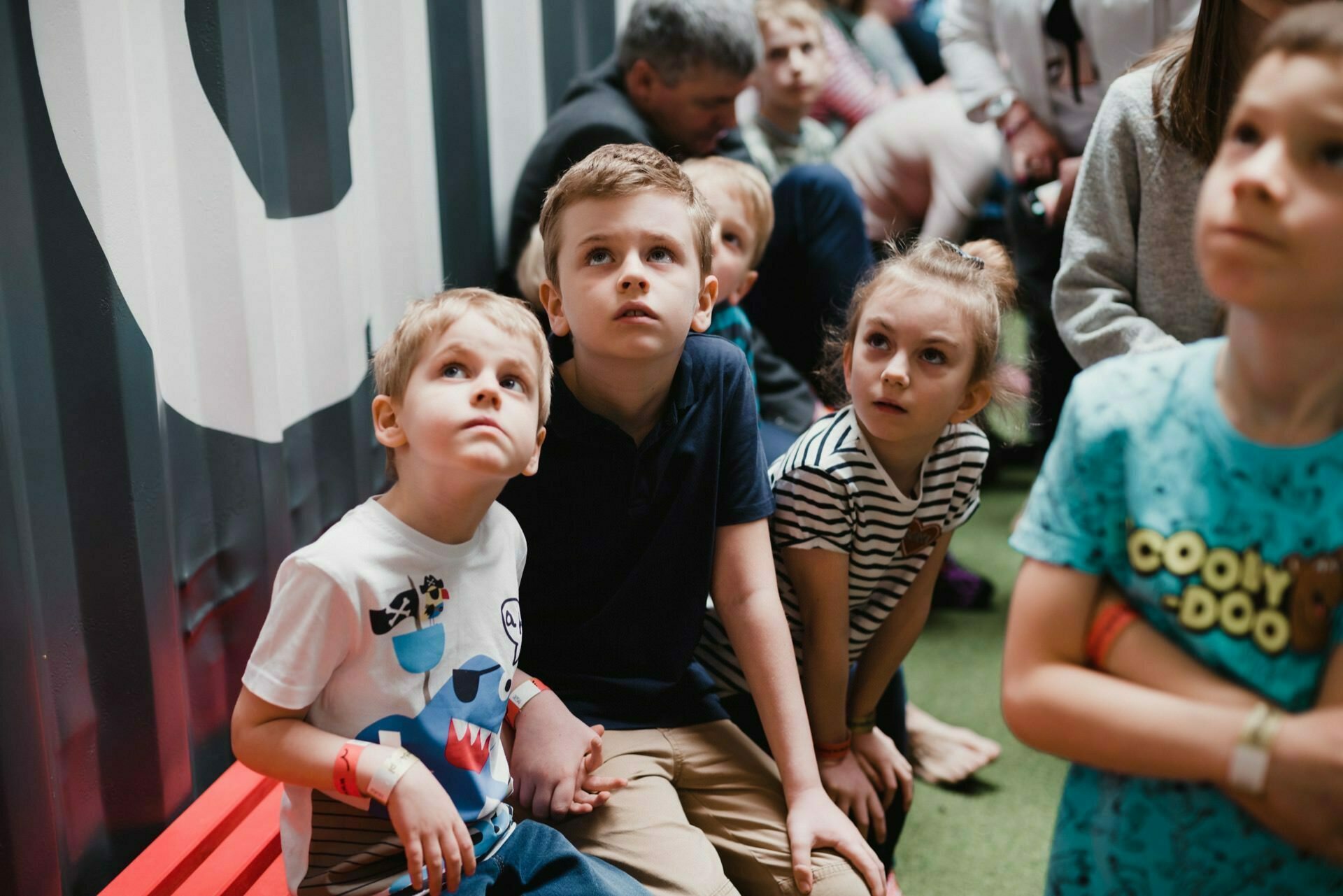 A group of children sit close together on a red bench and look up with an attentive face. They are in a room with gray and white walls. The children are wearing ordinary clothes, and adults can be seen in the background, also looking in the same direction. This moment was captured by an event photographer warsaw.   