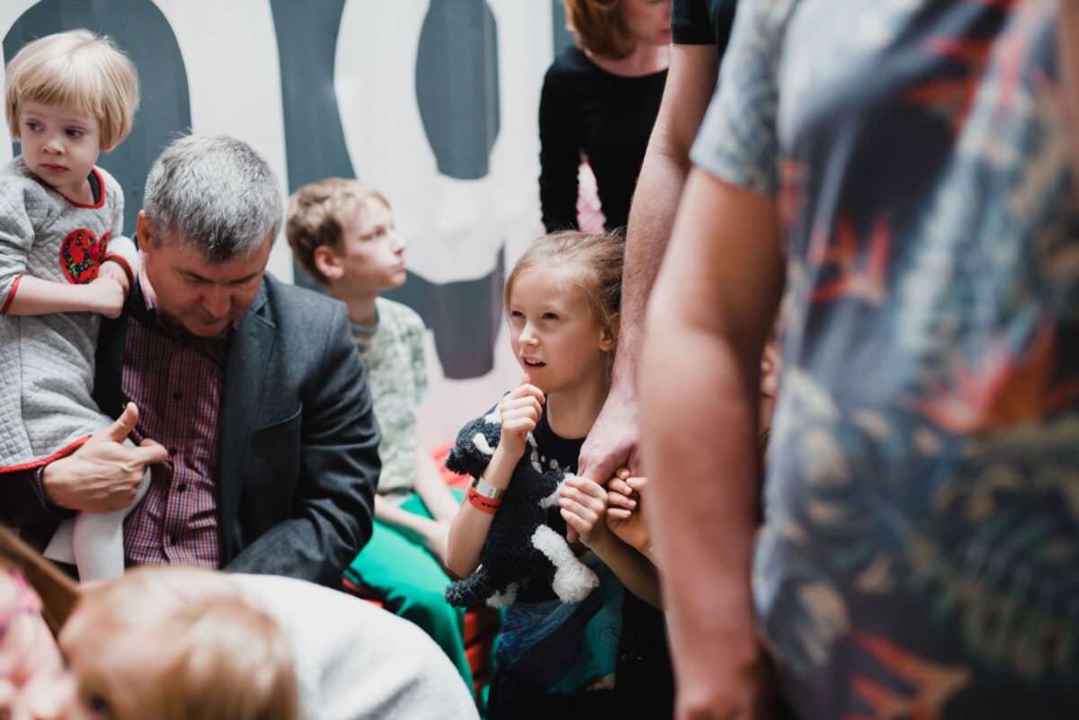 Grupa ludzi, w tym dzieci i dorośli, gromadzi się blisko siebie, tworząc tętniącą życiem scenę. Siwy mężczyzna trzyma małe dziecko w szarym swetrze, a dziewczyna z długimi włosami ściska pluszowego zwierzaka. Gwarna atmosfera oddaje istotę fotografii eventowej.