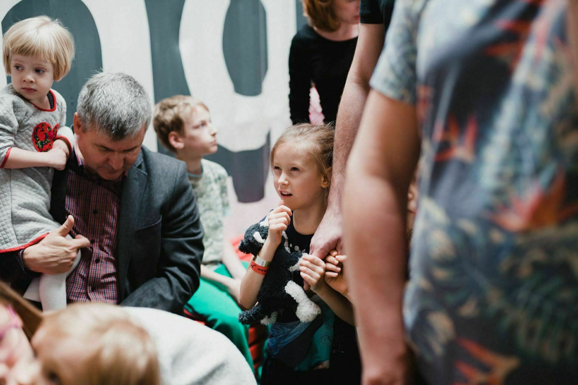 A group of people, including children and adults, gather close together, creating a vibrant scene. A gray-haired man holds a small child in a gray sweater, while a girl with long hair clutches a stuffed animal. The bustling atmosphere captures the essence of event photography.  