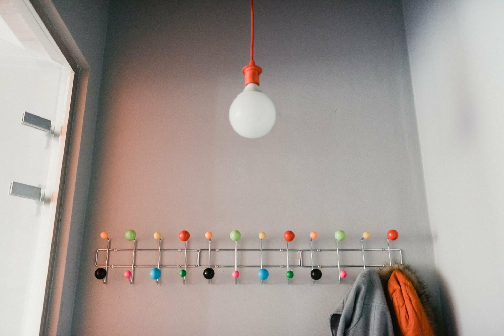 A room with a single hanging bulb fixture and a modern wall rack, reminiscent of a stylish studio event photographer warsaw. The rack has many colorful knobs, and coats can be hung on some of them. The wall is painted in a light gray color, and a partial glass door can be seen on the left side.  