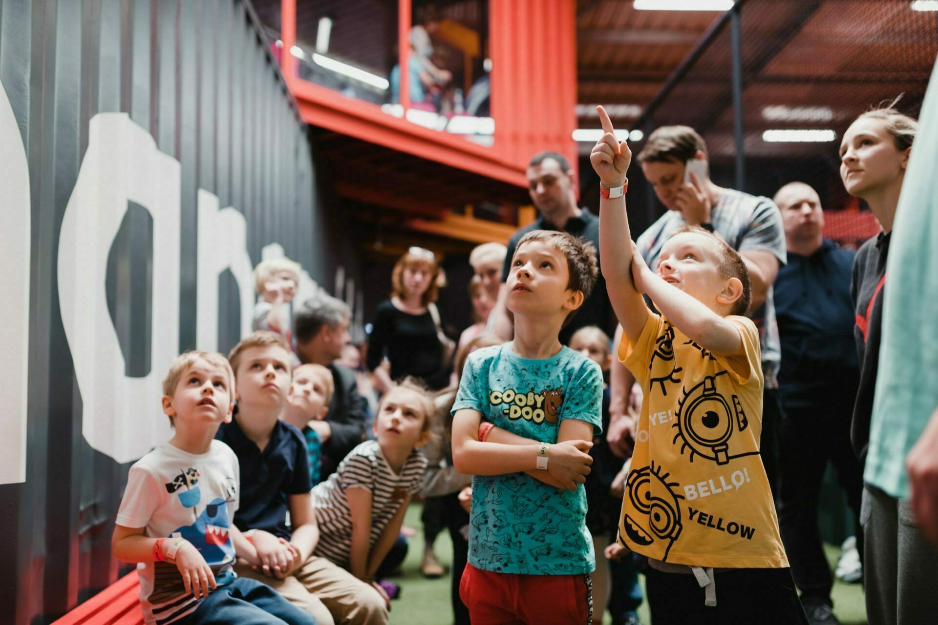 A group of children gather in a room and look up intently while one of them points to something out of frame. Some adults can be seen in the background. The environment suggests a fun or educational setting with vibrant colors and texture, beautifully captured in the event photography.  