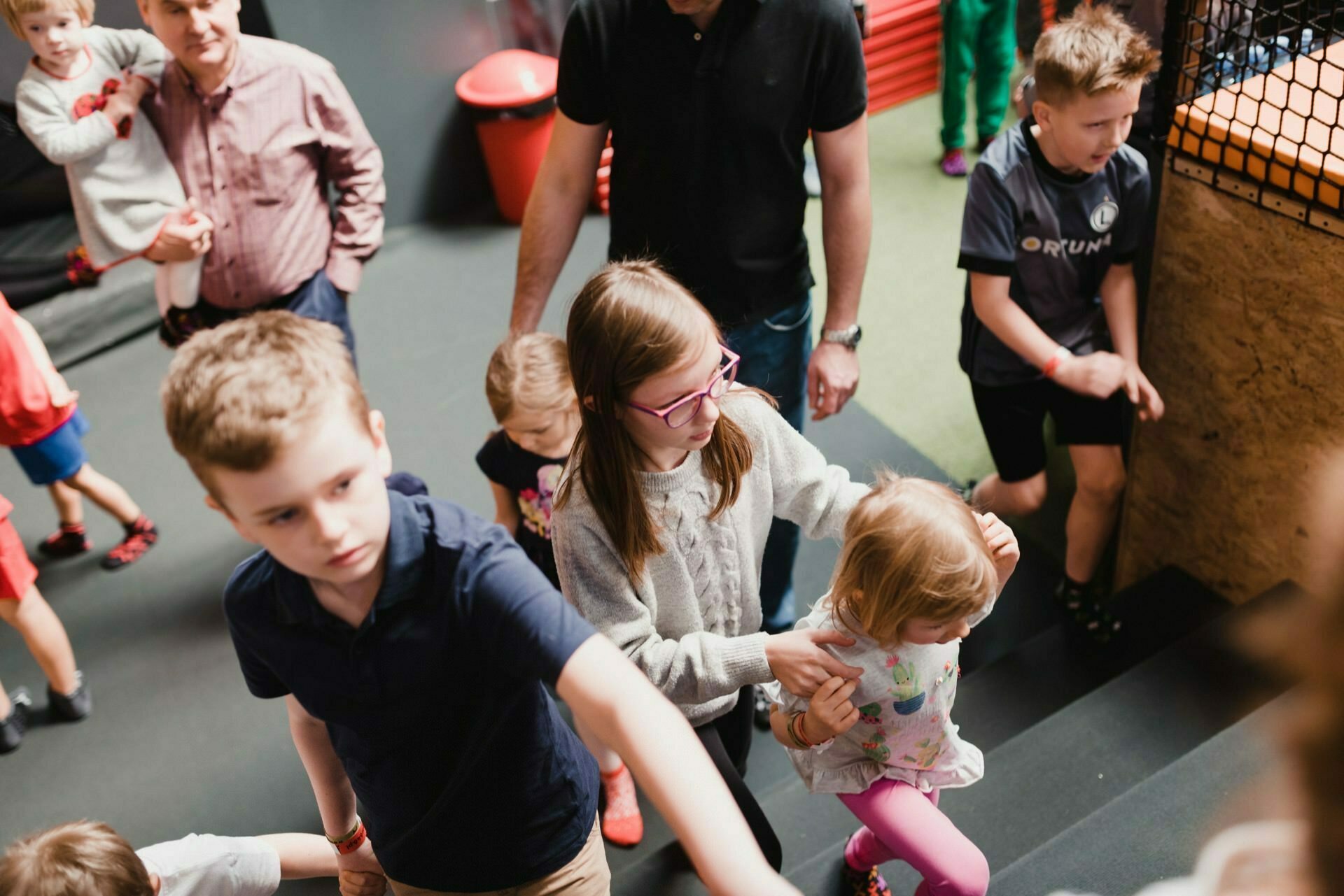 A group of children and adults walk up the stairs in a room. A woman wearing glasses helps a toddler climb the stairs, capturing the perfect moment for anyone interested in event photography. Other children and adults can also be seen, some holding hands and others climbing individually. The space looks busy and active.   