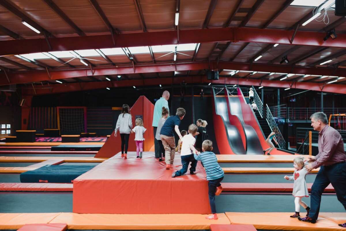 Grupa dorosłych i dzieci bawi się w krytym parku trampolin. Uchwyceni na tej fotografii eventowej, gromadzą się wokół czerwonej piankowej platformy ze zjeżdżalniami w tle. Obiekt idealny na fotorelację z wydarzeń wyposażony jest w trampoliny i inne urządzenia zabawowe pod wysokim stropem z metalowymi belkami.
