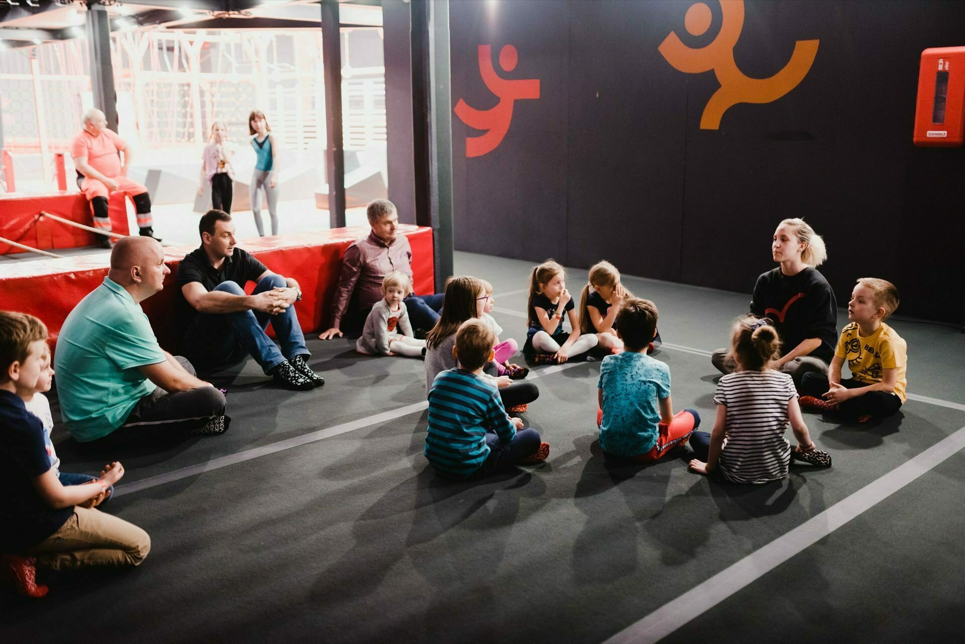 A group of children and adults sitting in a circle on the playground floor. The room, ideal for photo coverage of events, is decorated with fun wall graphics, and a trampoline is visible in the background. The scene looks like an organized activity or discussion.  