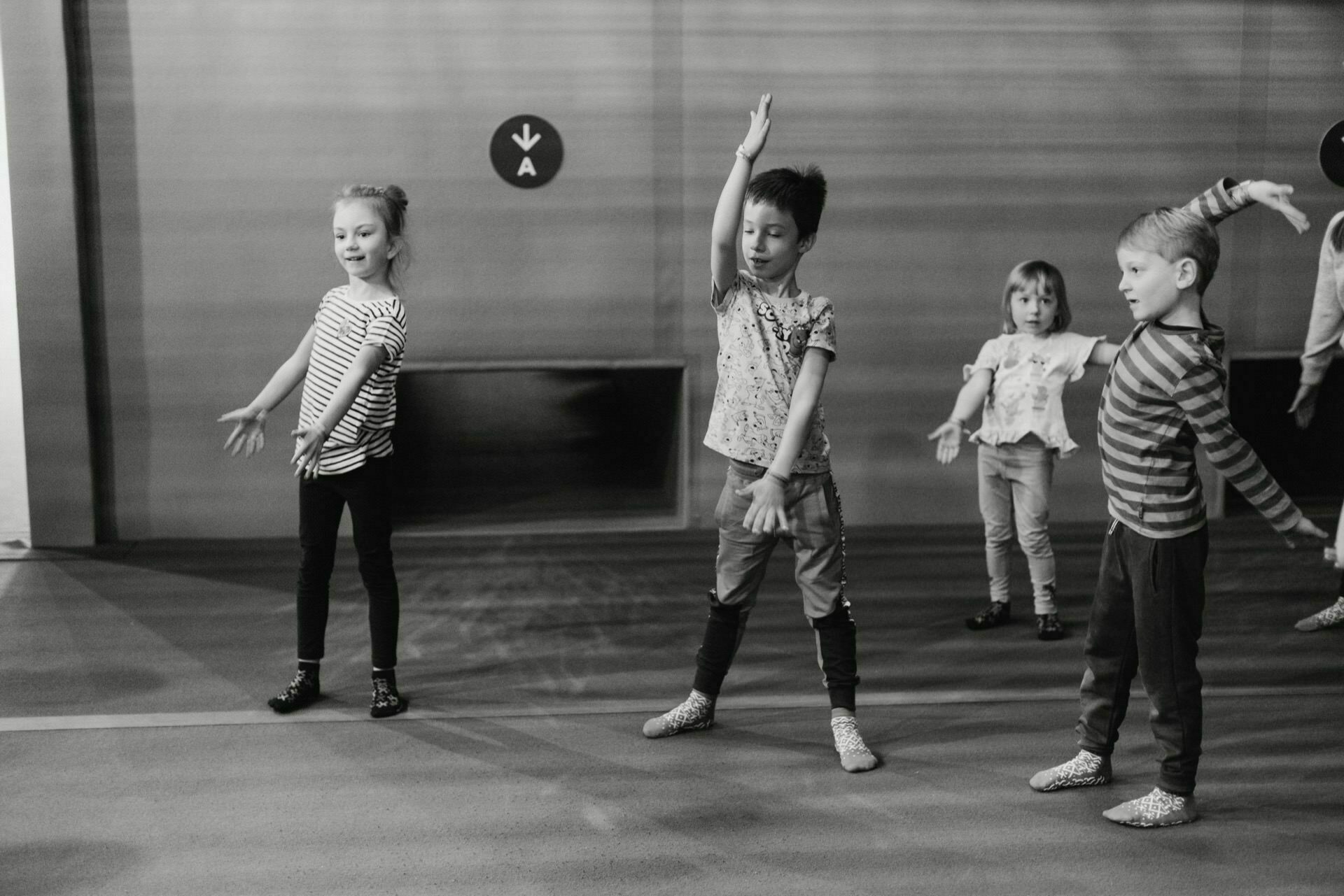 Black and white image of four children practicing dance moves in a studio. Each child stands with their hands raised in expressive poses, focusing on their movements. They are dressed in everyday clothes and seem to be enjoying the dance session - a beautifully captured captivating snapshot of the events.  