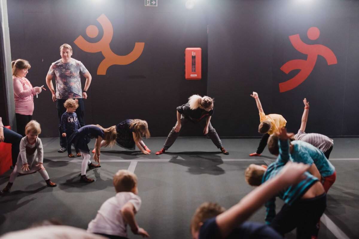 Grupa dzieci i nauczycielka rozciągają się na sali gimnastycznej, co pięknie uchwyciła fotografia eventowa. Dzieci pochylają się, aby dotknąć palców u nóg, podczas gdy nauczyciel demonstruje rozciąganie. Czarne ściany sali gimnastycznej ozdobione stylizowanymi postaciami w kolorze czerwonym i pomarańczowym tworzą żywe tło, z gaśnicą zamontowaną na ścianie.