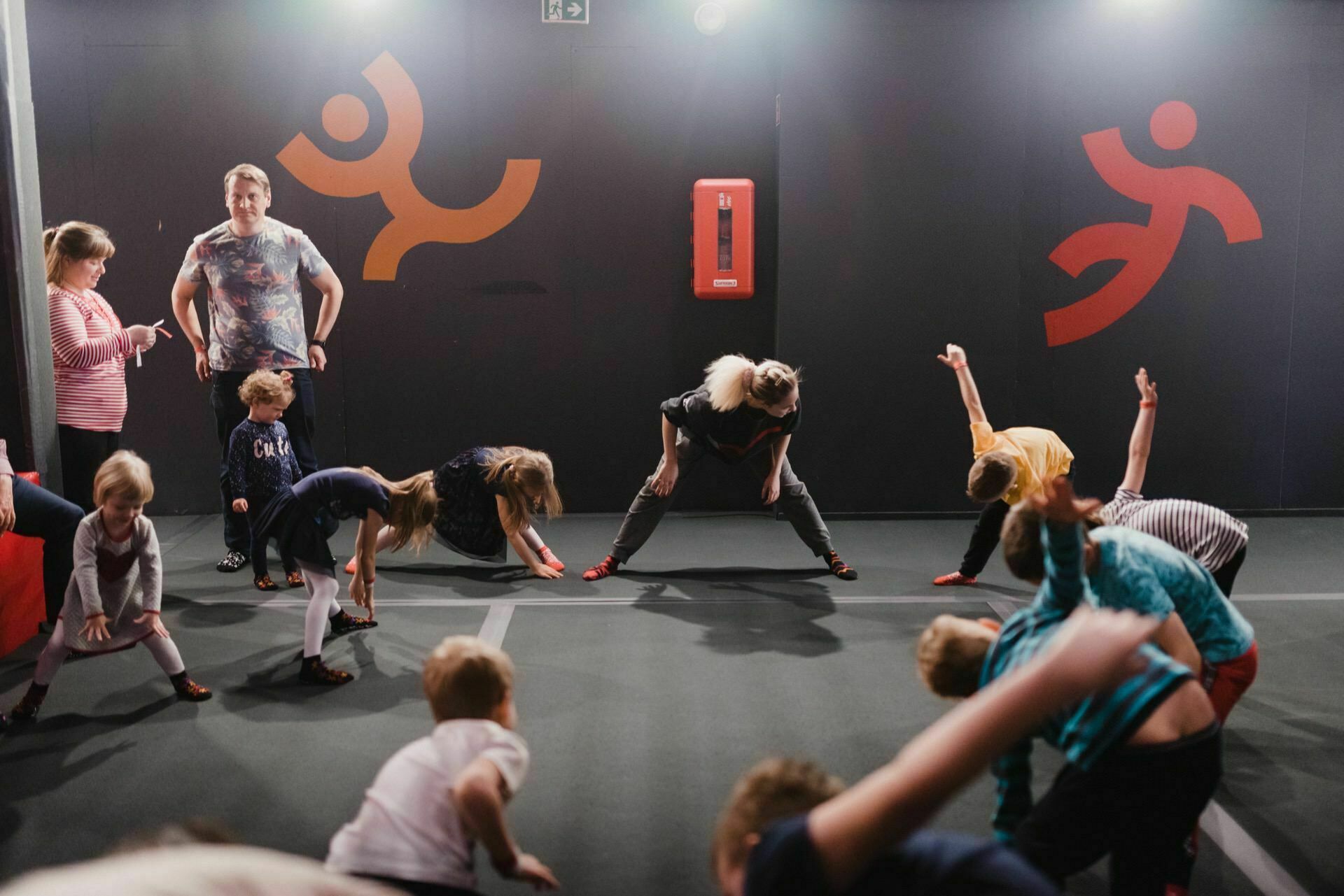 A group of children and a teacher are stretching in a gymnasium, as beautifully captured by an event photo. The children bend over to touch their toes while the teacher demonstrates stretching. The black walls of the gymnasium decorated with stylized figures in red and orange create a lively backdrop, with a fire extinguisher mounted on the wall.  