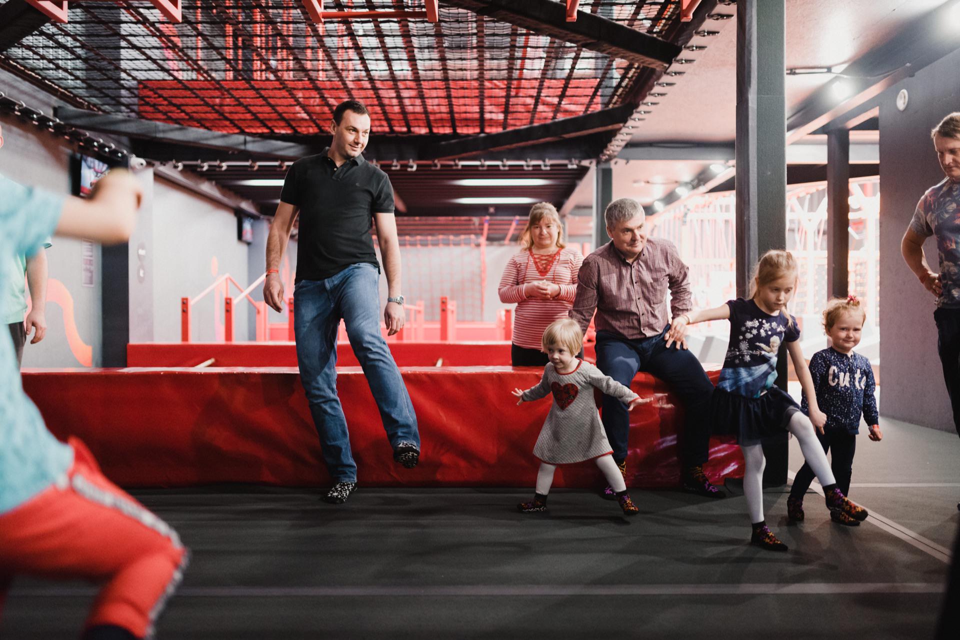 A group of adults and children play together in a room. The scene is vibrant with red accents and playground equipment in the background. Everyone seems engaged and smiling, which is reminiscent of a photo report of events in a fun and energetic environment such as a playground or entertainment center.  