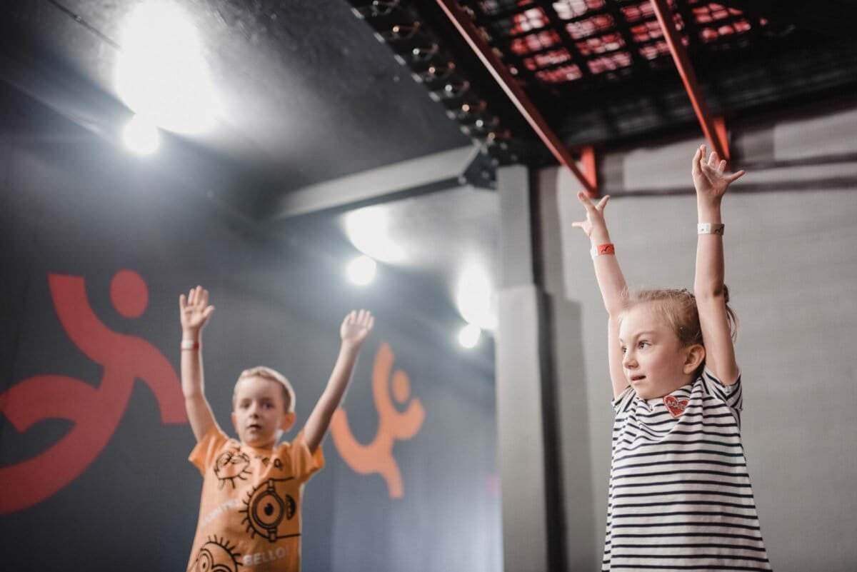 Dwoje dzieci z podniesionymi rękami uczestniczy w zajęciach w pomieszczeniu. Dziecko po lewej stronie ma na sobie pomarańczową koszulę z czarnymi wzorami, a dziecko po prawej stronie nosi koszulę w paski. Tło jest szare z abstrakcyjnymi pomarańczowymi postaciami na ścianie i drążkami nad głową, doskonale uchwyconymi na tej fotografii eventowej.