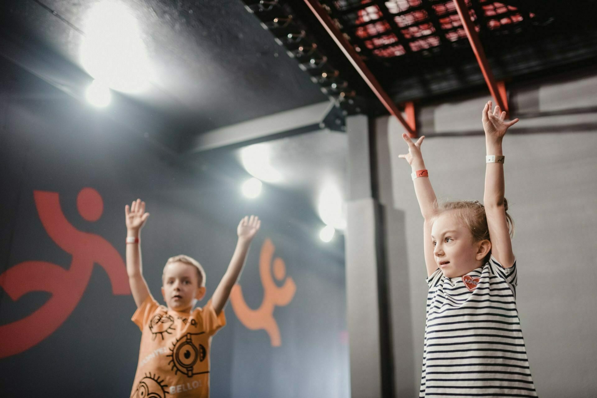 Two children with their hands raised participate in an activity in a room. The child on the left is wearing an orange shirt with black patterns, and the child on the right is wearing a striped shirt. The background is gray with abstract orange figures on the wall and rods overhead, perfectly captured in this event photograph.  