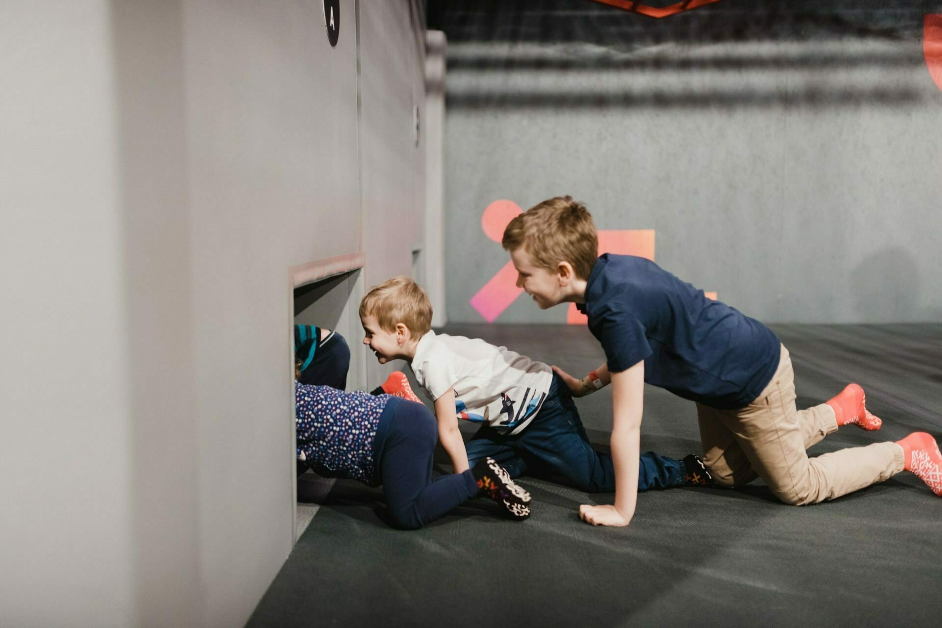 Three children are crawling on the floor and peering into a small rectangular opening in a gray wall. The children are dressed in plain clothes, and the scene appears to be part of an interactive exhibition or play environment, perfect for a photo essay of events captured by an event photographer Warsaw. 