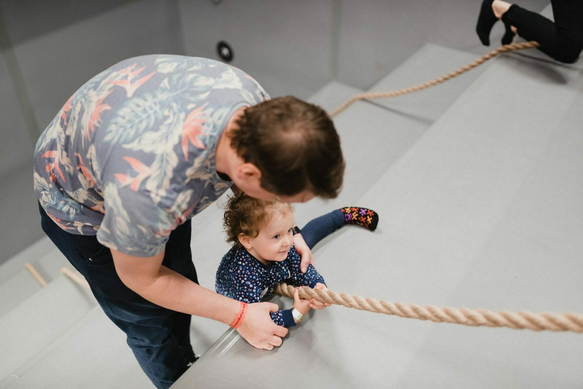 A man helps a young child climb a gray sloping surface using a rope. A man dressed in a colorful floral shirt holds a focused child holding a rope. In the background, a second person, partially visible, is climbing. This moment perfectly captures the essence of event photography.   