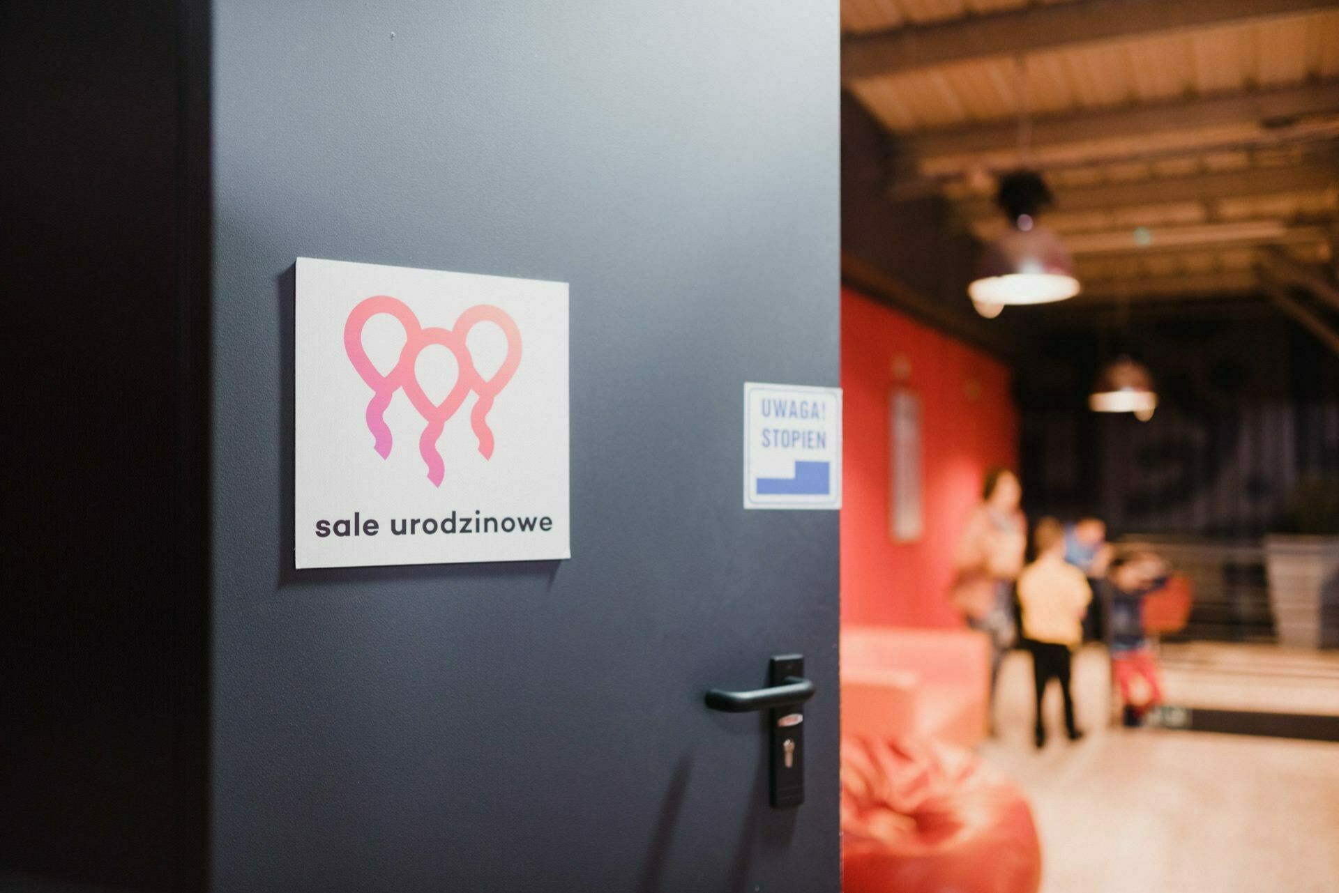 The dark door is partially open, revealing a sign with three pink balloon icons and the words "birthday sale" (birthday rooms). In the room, vague people appear to be socializing near red couches and a modern industrial-style interior - perfect for an event photo shoot. 