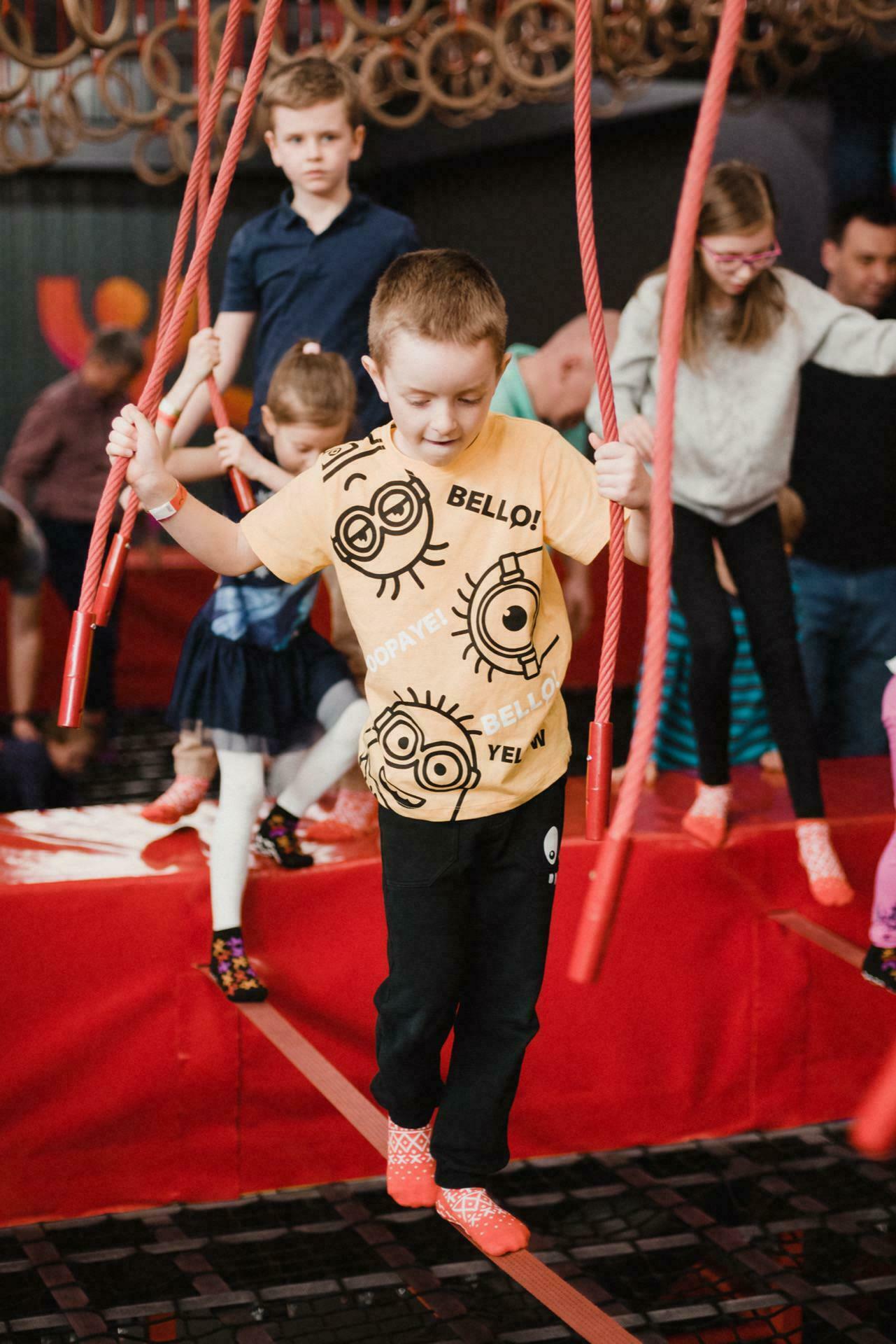 Children play on a rope obstacle course in a room. A boy in a yellow shirt with cartoon faces holds onto red ropes while balancing on a beam. Other children in colorful costumes wait their turn or traverse the track in the background, perfect for an event photo shoot.  