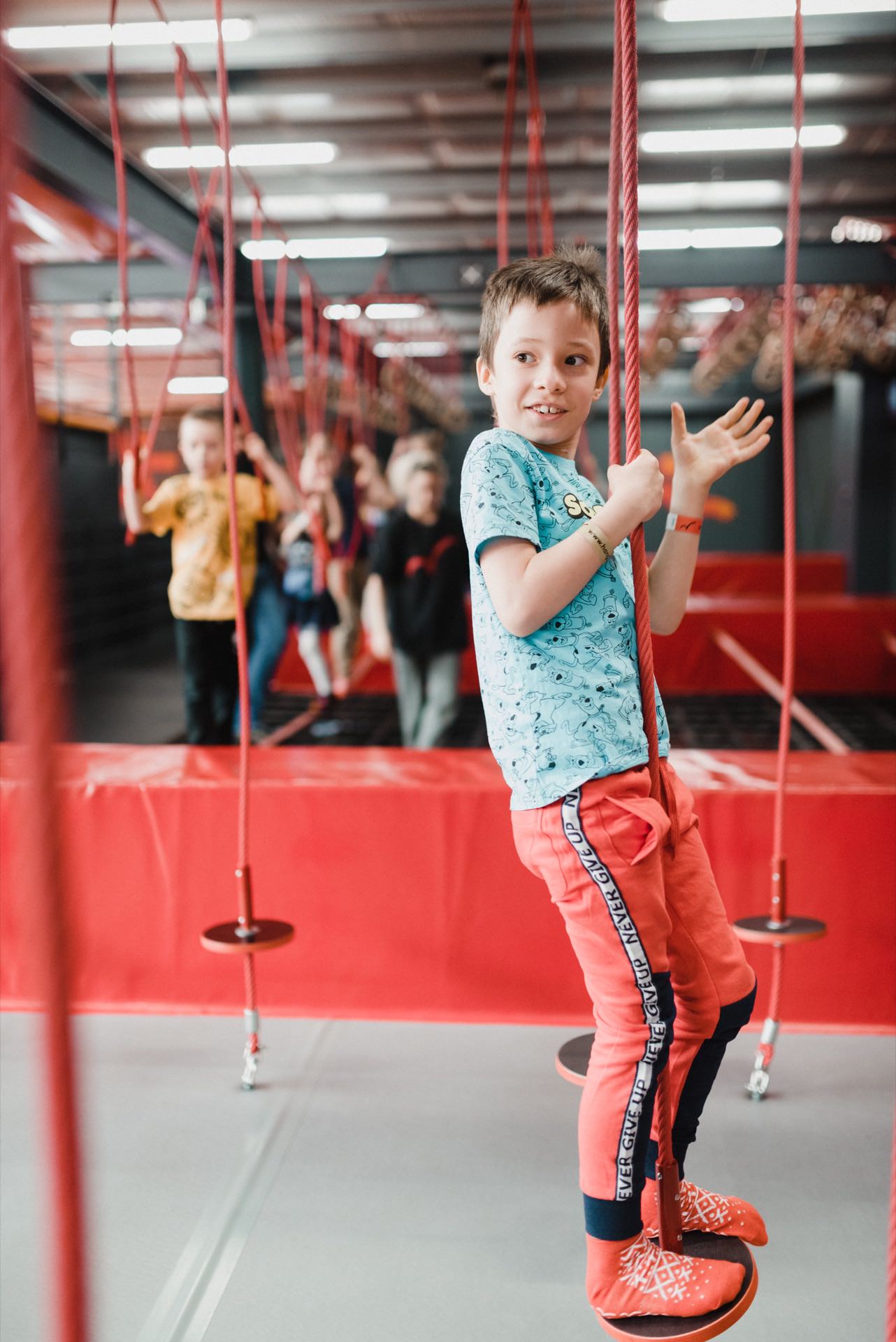 A young boy in a blue shirt and red pants holds a red rope while balancing on a platform in an indoor playground. He smiles and looks to the side, captured perfectly for event photography. Several other children in the background appear to be playing on similar equipment.  