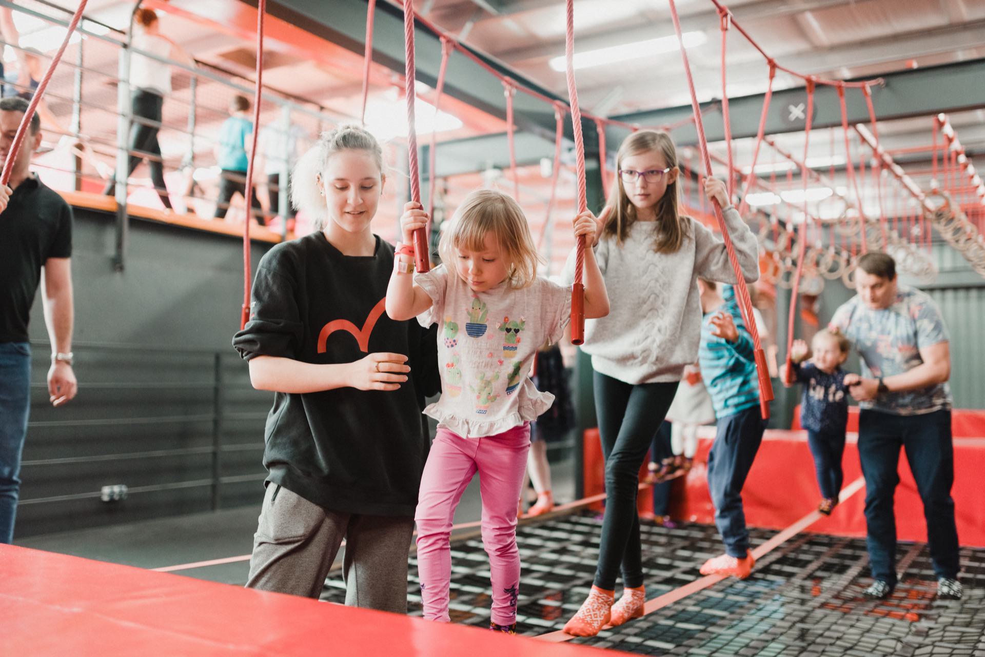 A young girl walks on a rope bridge over an indoor obstacle course, accompanied by an older girl. Other children and adults also participate in the background. In a vibrant and energetic environment, captured in a delightful photo essay of the events, red and black dominate the decor.  