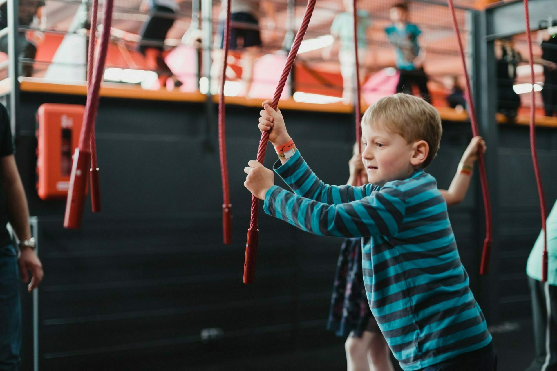 A young boy in a blue striped shirt plays with hanging red ropes in an indoor activity center. Other children are visible in the background, participating in similar activities. The environment looks fun and engaging with the various play structures, perfectly capturing the lively photo-reportage of the events.  