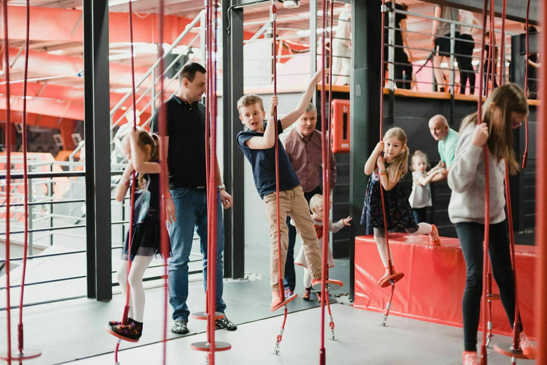 A group of children and adults participate in a rope obstacle course activity in a room with red and black structures. The children balance on the ropes while the adults supervise and assist. The atmosphere is lively and energetic, perfect for capturing photo reports of the events.  