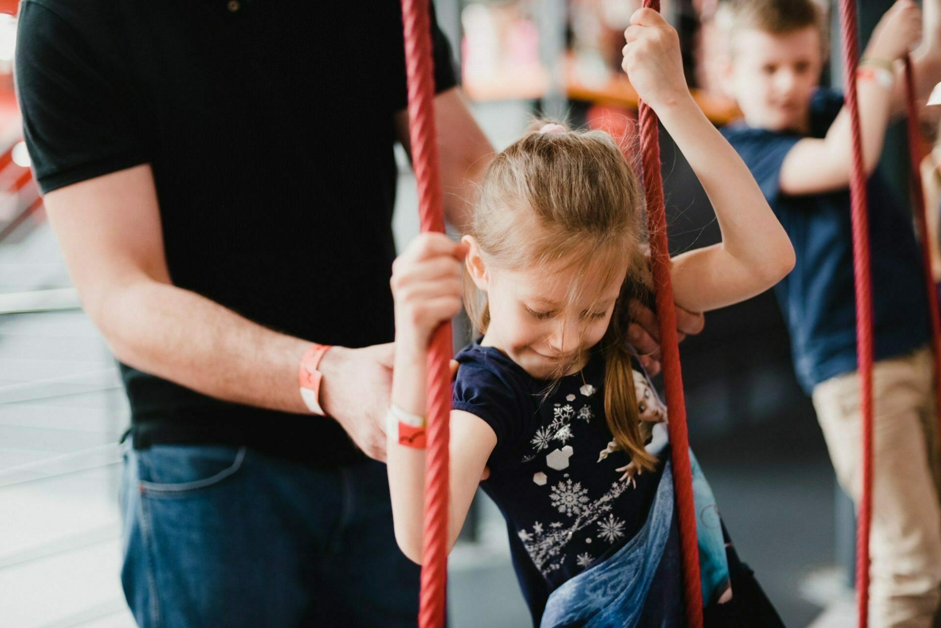 A young girl with pigtails holds two red ropes, while a man in a black shirt and jeans stands next to her, offering support. Another child can be seen in the background. The scene, ideal for event photography, appears to be set in an indoor playground or activity center.  