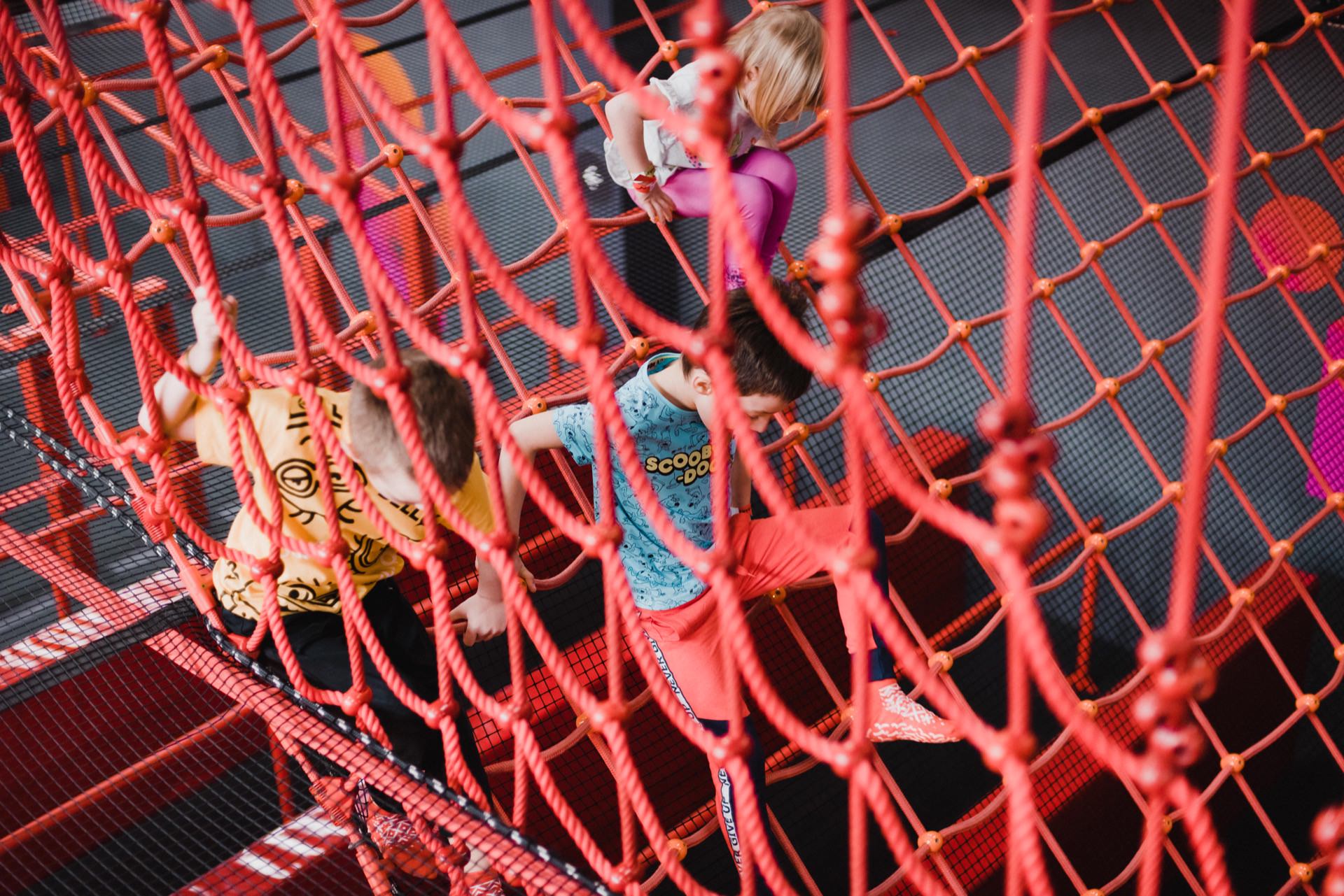 Children playing on a large red rope climbing structure in an indoor playground have been captured in a vibrant event photo essay warsaw. They move along the intertwined ropes, looking focused and enthusiastic. The background shows a padded floor for safety and a vibrant playground environment.  