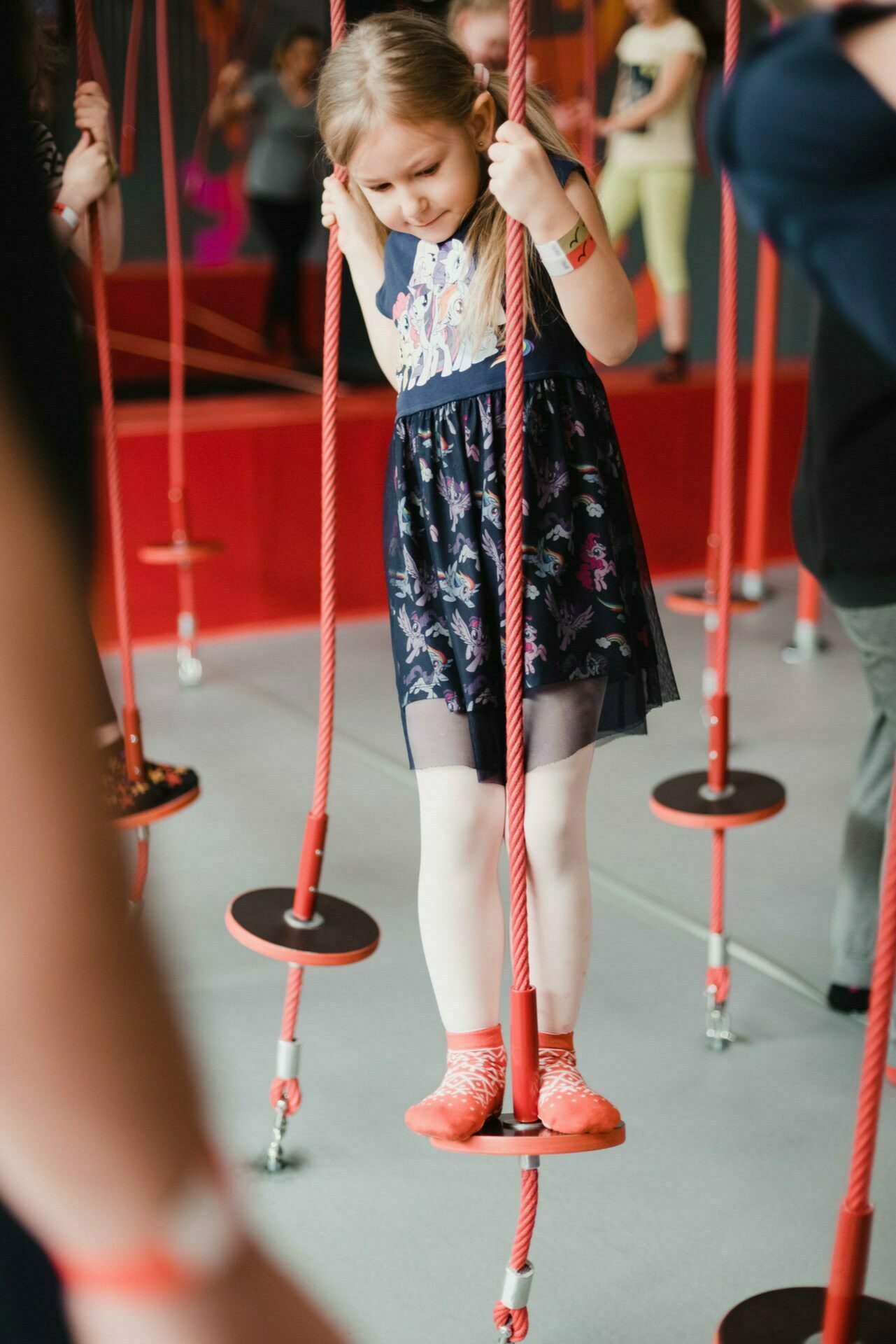 A young girl in a dress balances carefully on hanging platforms connected by ropes in an indoor playground. She holds onto the ropes with both hands and looks focused on her steps. Other people, also involved in the action, are visible in the background, making it an ideal scene for an event photographer Warsaw.  