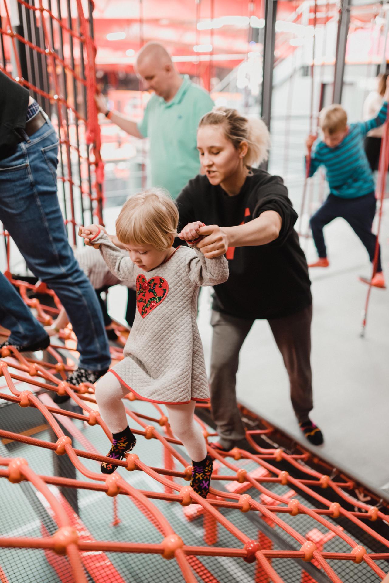 A small child dressed in a gray dress with a heart motif walks through the net, which was perfectly captured by an event photographer Warsaw. An adult woman helps by holding her hands. Other people in the background are also moving through the net climbing area, contributing to the lively and fun atmosphere of this event photo shoot.  