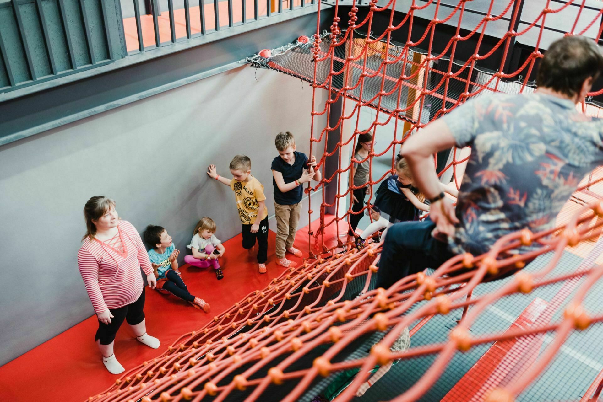 A group of children and a few adults are actively participating in activities at an indoor playground. Some children are climbing on the red net, while others are sitting or standing by the gray wall. The ground has a bright red cushioning surface, and the area appears lively and playful, perfect for photo coverage of events.  