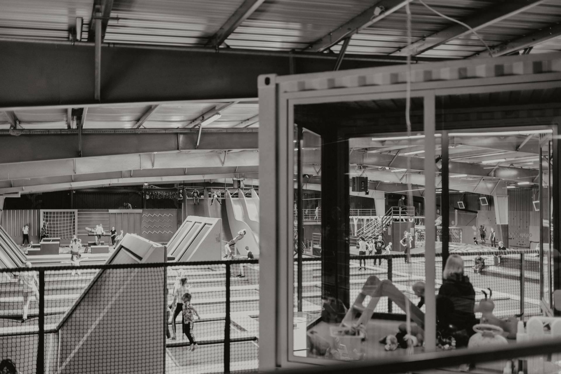 A black-and-white image of an indoor trampoline park captures a dynamic scene: people jumping on trampolines, climbing on structures. In the foreground, a glassed-in area provides a view of what's happening below, with some people casually observing the situation from inside. This is a perfect example of event photography in action.  