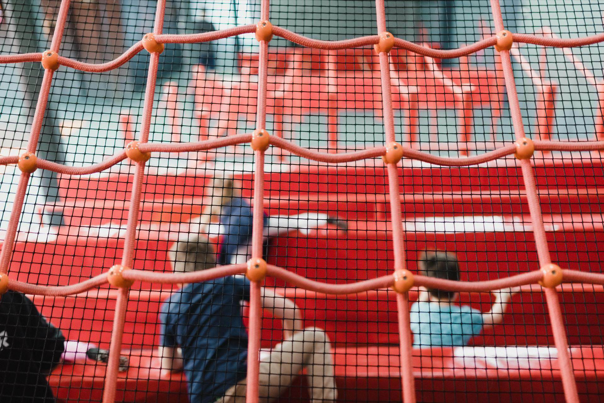 Several children are playing and climbing the bright red stepped structures, seen from the foreground through the grid. The black and orange grid-like pattern partially obscures their activity in this bustling indoor playground - the perfect photo for any event photography enthusiast or event photographer Warsaw. 