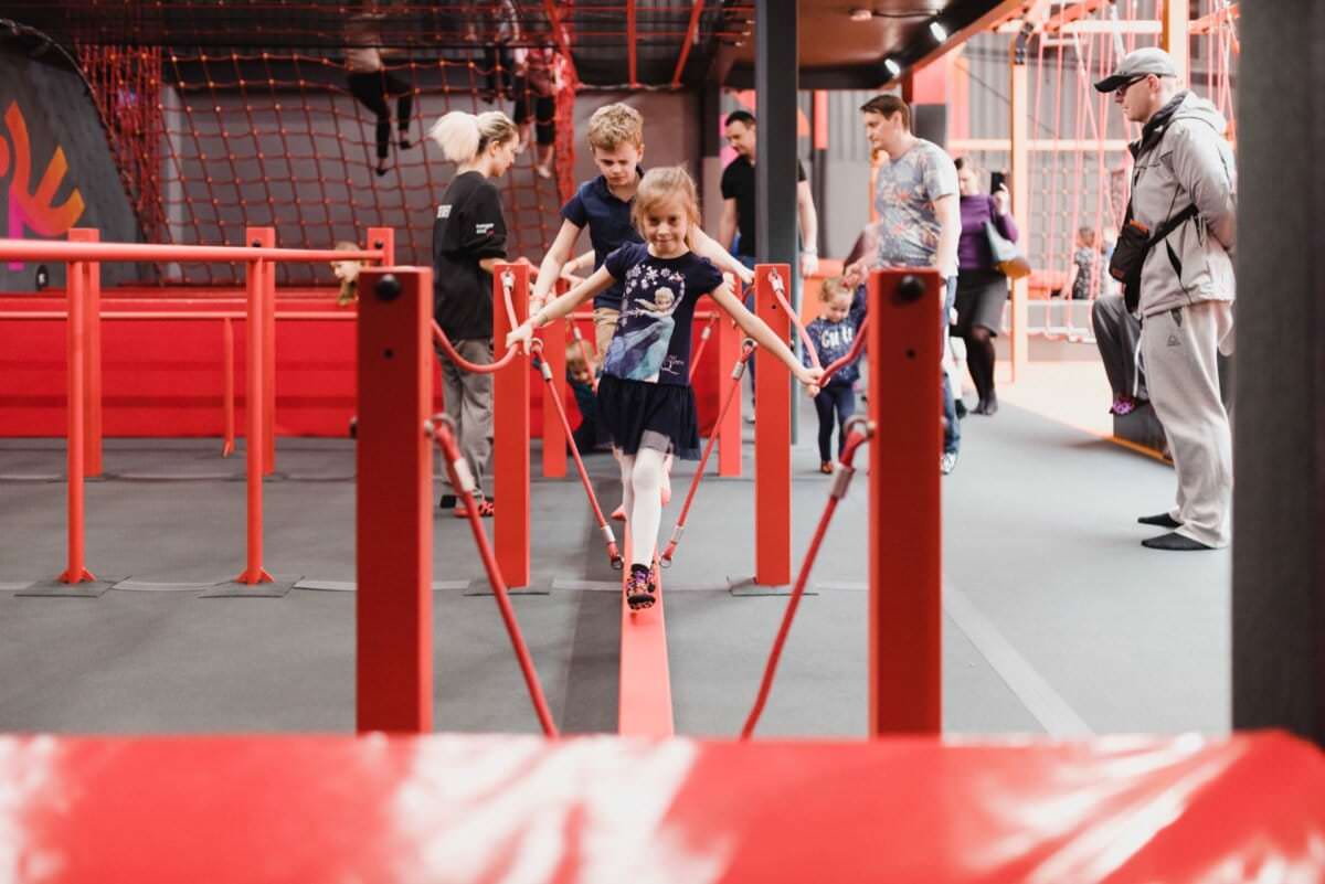 Młoda dziewczyna balansuje na czerwonej belce w pomieszczeniu centrum zabaw, otoczona innymi osobami, w tym chłopcem i kilkoma dorosłymi. Środowisko może pochwalić się czerwonymi i szarymi strukturami, siatkami i sprzętem przypominającym salę gimnastyczną. Żywa atmosfera sprawia wrażenie, jakby ktoś miał *fotografię na ścianie przyszłej*, uwieczniając każdą radosną chwilę.